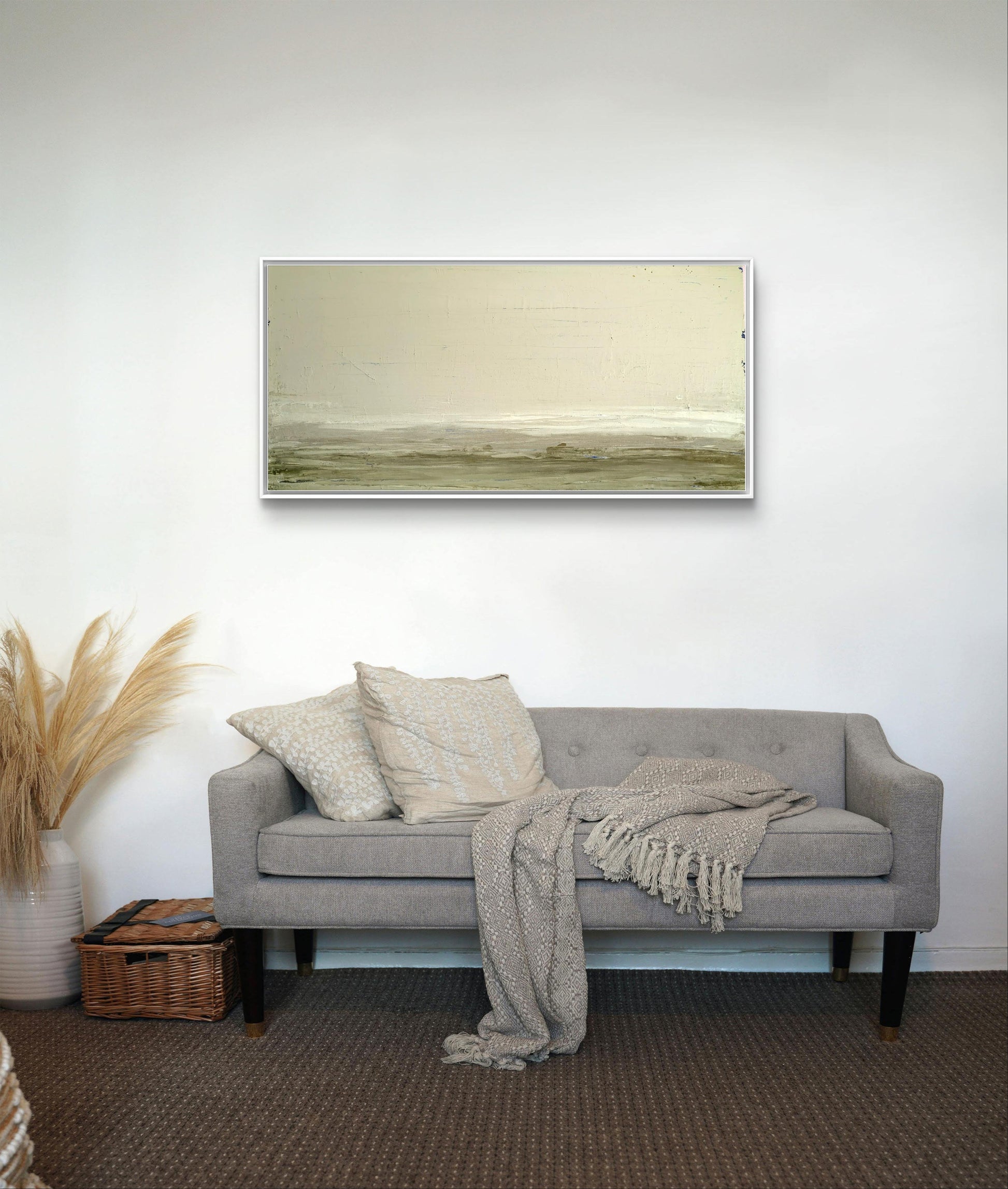 Ein hellgraues Sofa mit strukturierten Kissen und einer Strickdecke steht vor einer weißen Wand, darunter "Zwischen Sommer und Herbst II" von Conny Wachsmann. Ein Korb und Pampasgras-Dekor stehen links auf einem braunen Teppichboden.