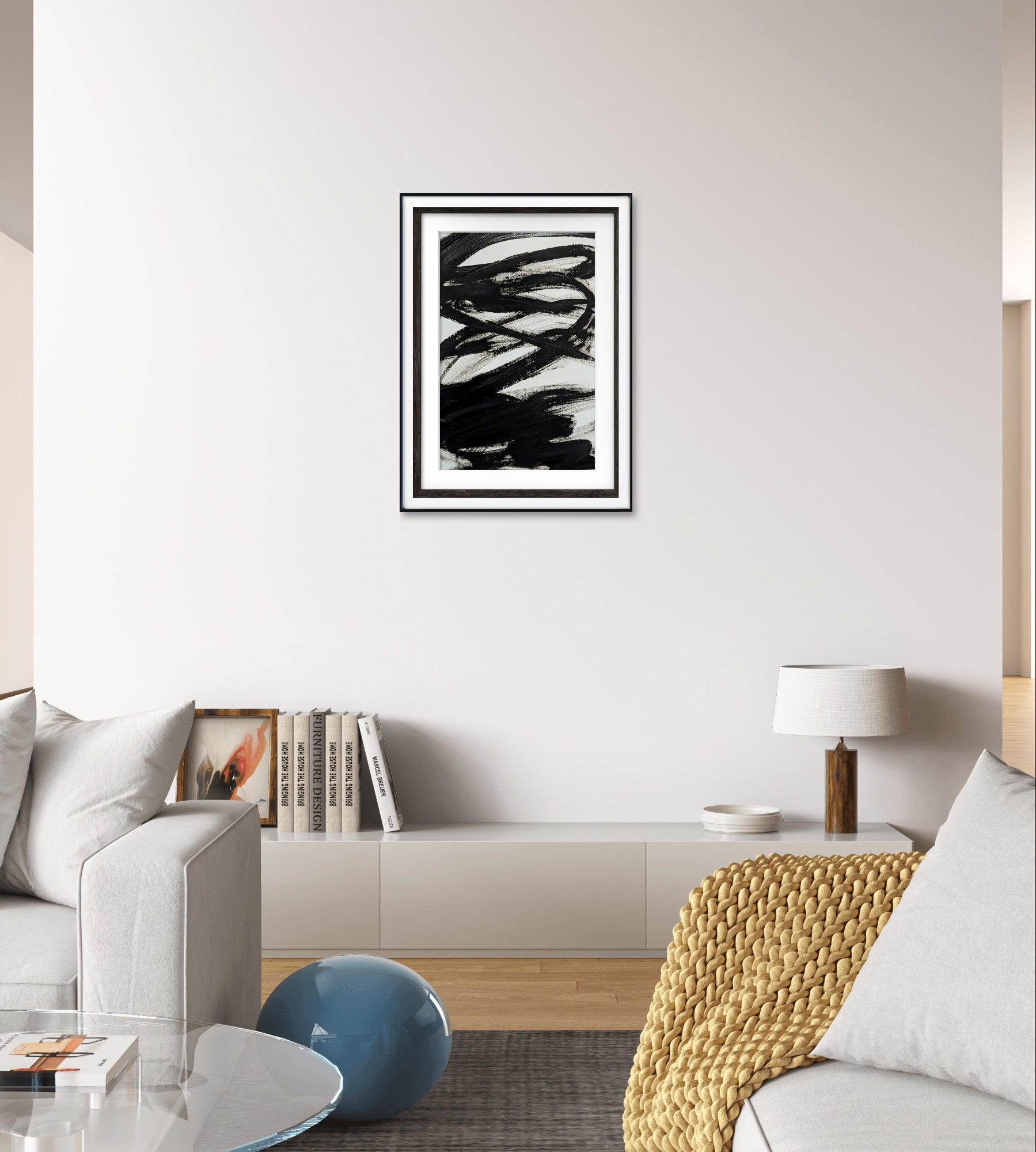 Ein modernes Wohnzimmer mit einem weißen Sofa, einer strukturierten beigen Decke und einer minimalistischen weißen Lampe. "Zeichnungen & Reduktion III" von Conny Wachsmann, Künstlerin, hängt über einem niedrigen Regal, das mit Büchern und Dekoartikeln gefüllt ist.