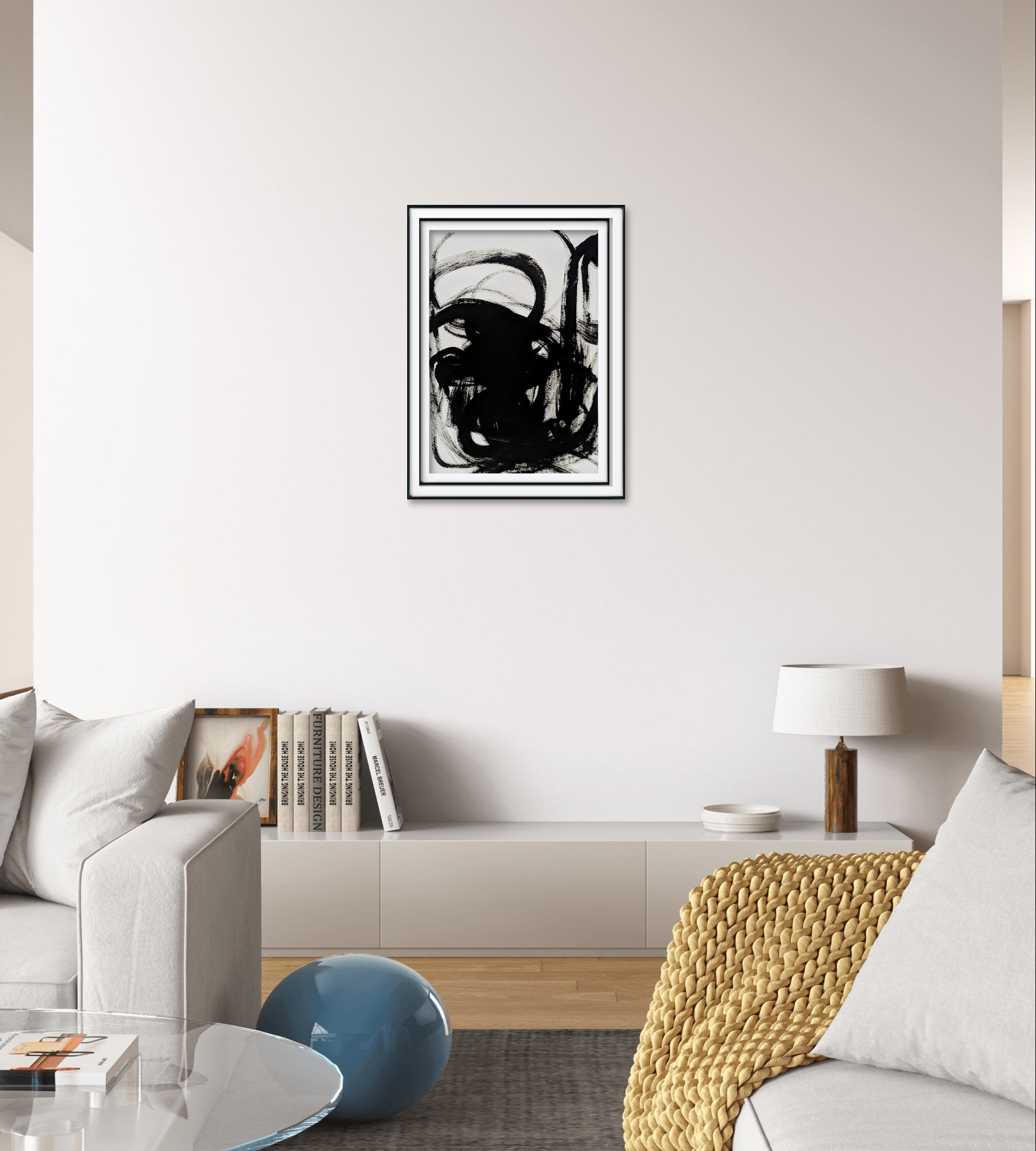 Ein modernes Wohnzimmer mit einem grauen Sofa, einem niedrigen weißen Schrank, einer Tischlampe, Büchern, einem blauen runden Dekostück und "Zeichnungen & Reduktion IV" von Conny Wachsmann - eine minimalistische Schwarz-Weiß-Zeichnung in einem schmalen Rahmen an der weißen Wand.