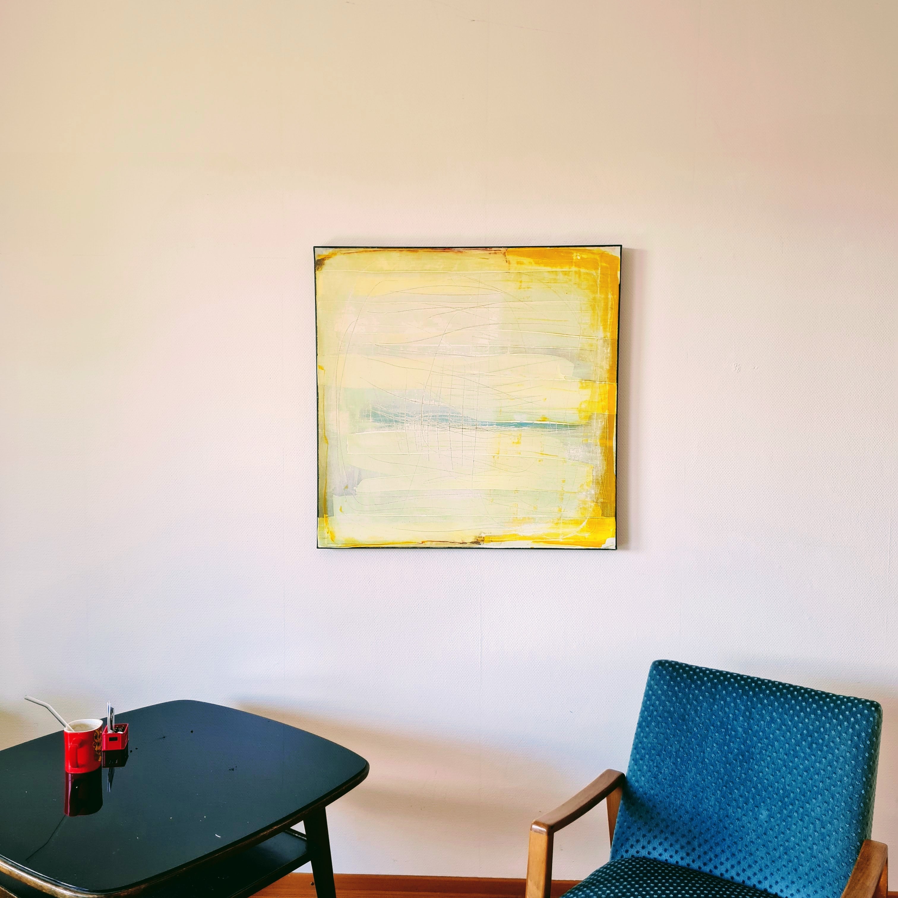 Küstenzauber II von Conny Wachsmann, Künstlerin, eine gerahmte abstrakte Landschaft in Gelb- und Hellblautönen, hängt an einer weißen Wand über einem blauen Stuhl. Ein schwarzer Tisch daneben mit einer roten Tasse und einem Löffel unterstreicht das minimalistische, moderne Dekor.