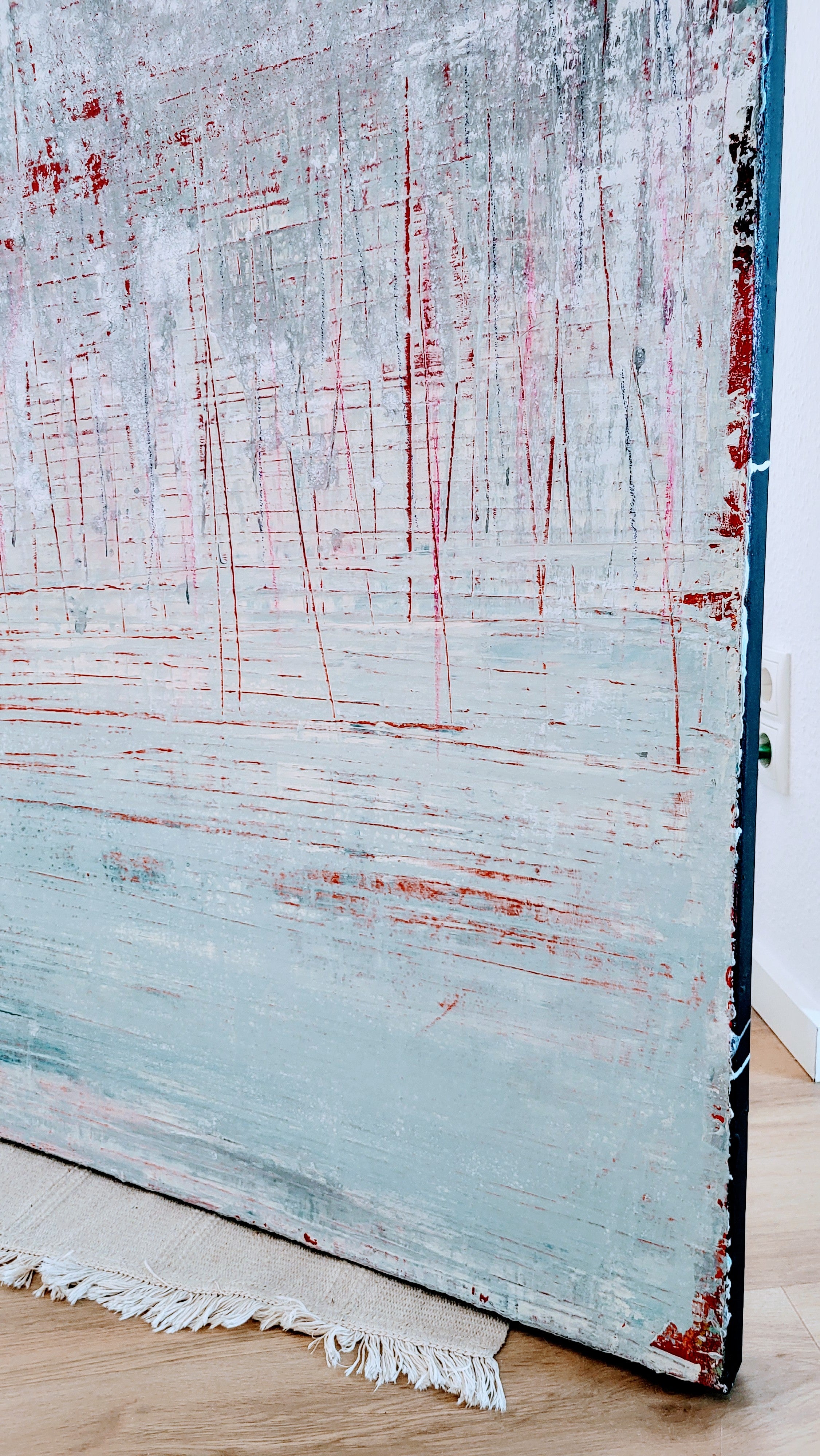 Vorschiedene Weiten" von Conny Wachsmann, ein abstraktes blau-weißes Gemälde mit vertikalen roten und weißen Streifen in Kratztechnik, steht an einer Wand auf einem Fransenteppich über einem Holzboden - ideal für jede Kunstgalerie.