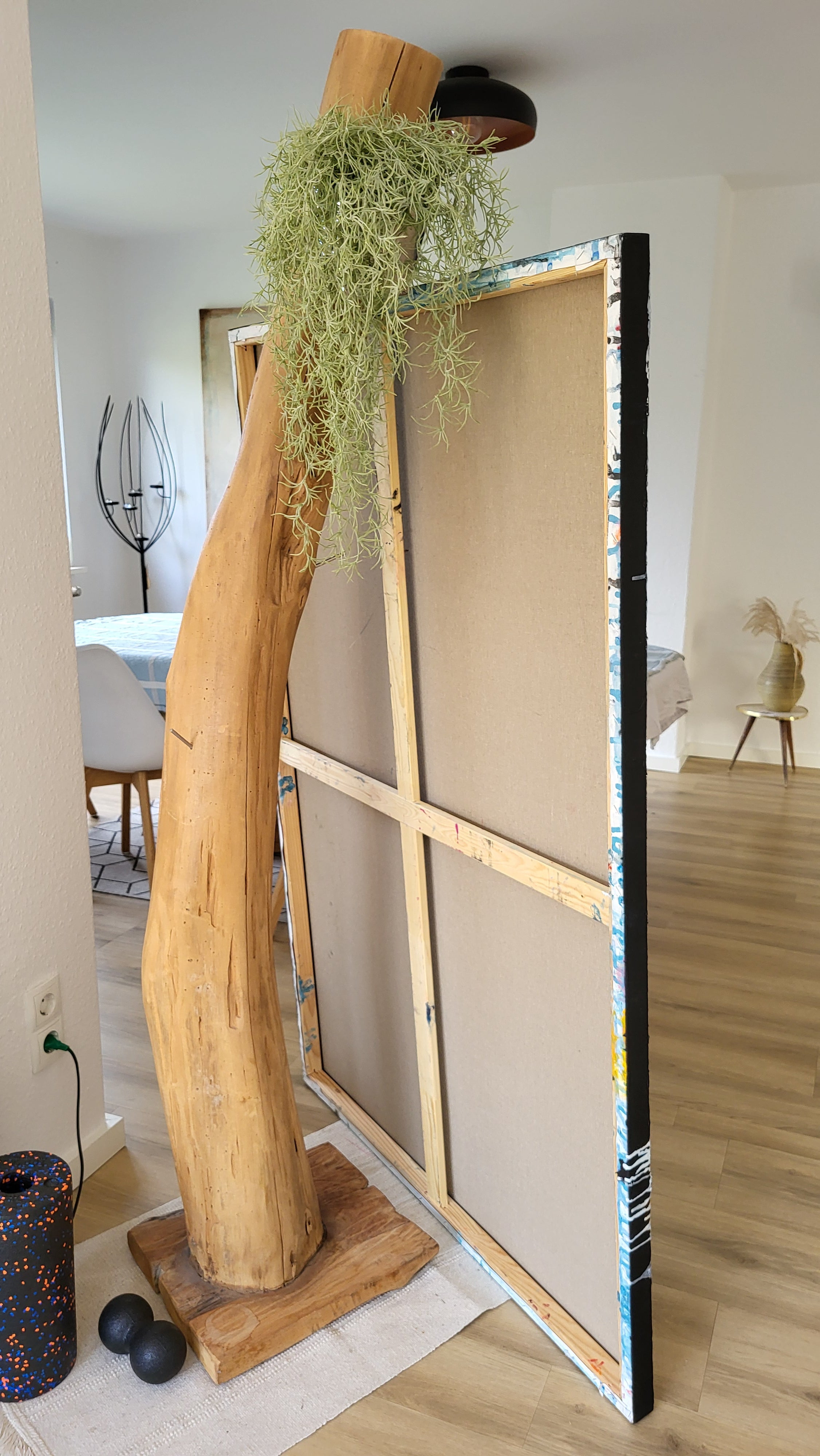 Vorschiedene Weiten", ein Werk der Künstlerin Conny Wachsmann, zeigt einen großen Baumstamm mit einer Pflanze, der an der Rückseite einer hohen, gerahmten Leinwand auf einem Holzsockel befestigt ist und an den modernen Galeriestil erinnert.