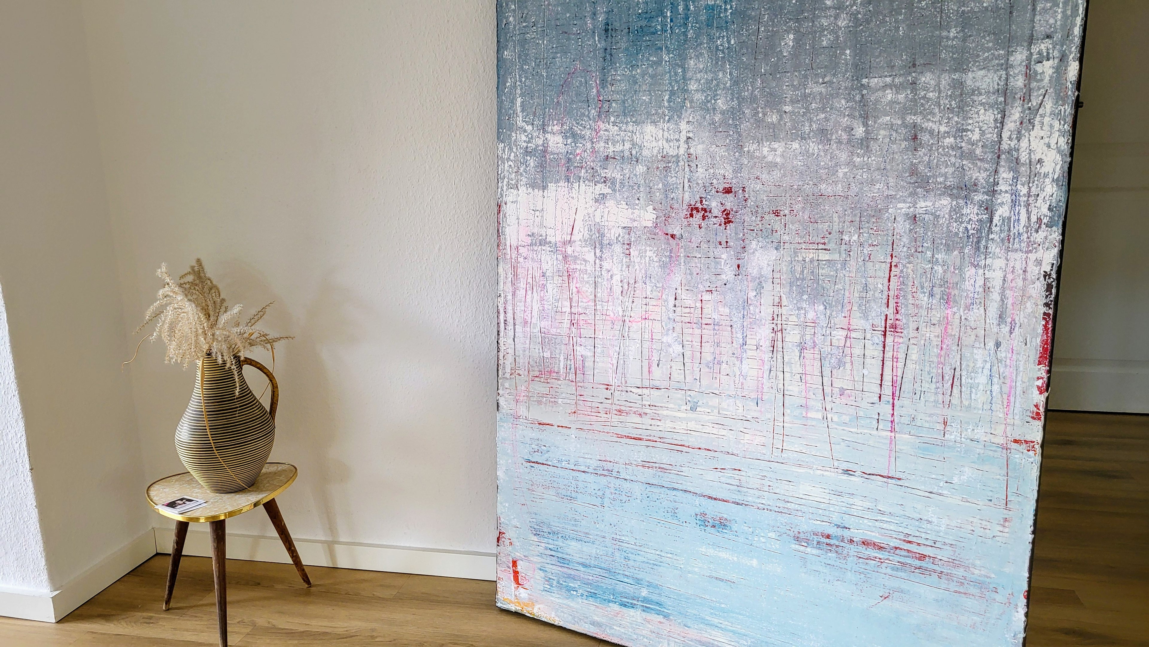 Ein großes Kunstwerk mit dem Titel "Vorschiedene Weiten" von Conny Wachsmann, Künstlerin, das in Zusammenarbeit mit Kratztechnik entstanden ist, lehnt an einer weißen Wand neben einer gestreiften Keramikvase auf einem runden Tisch und erinnert an den Stil einer artvergnuegen galerie.