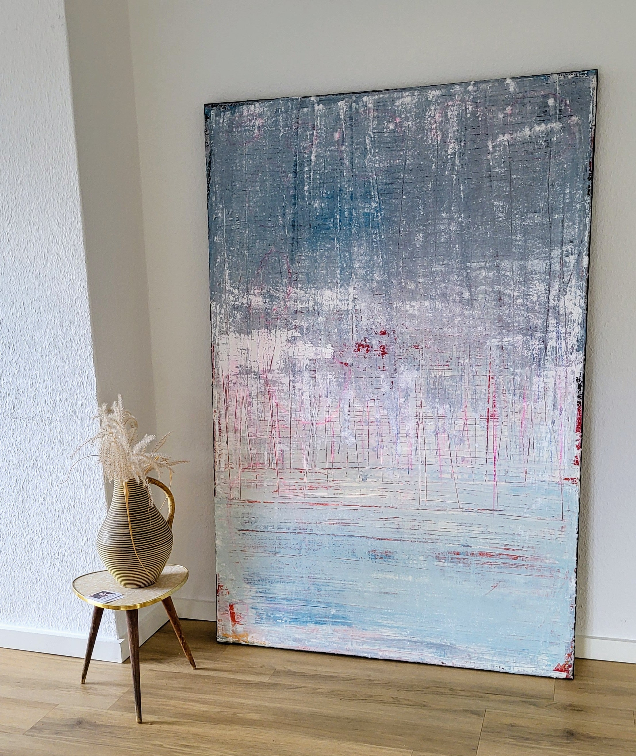 An einer weißen Wand lehnt das großformatige Werk "Vorschiedene Weiten" von Conny Wachsmann. Daneben steht auf einer Holzschemel eine Keramikvase mit Gräsern - ein stilvoller Look wie aus der artvergnuegen Galerie.