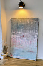 Vorschiedene Weiten von Conny Wachsmann, Künstlerin - ein großes abstraktes Bild in Pastellblau, Rosa und Weiß - lehnt vor einer weißen Wand unter Schwarzlicht. Ein kleiner Hocker mit getrockneten Gräsern vervollständigt die galerieartige Auslage auf dem Holzboden.