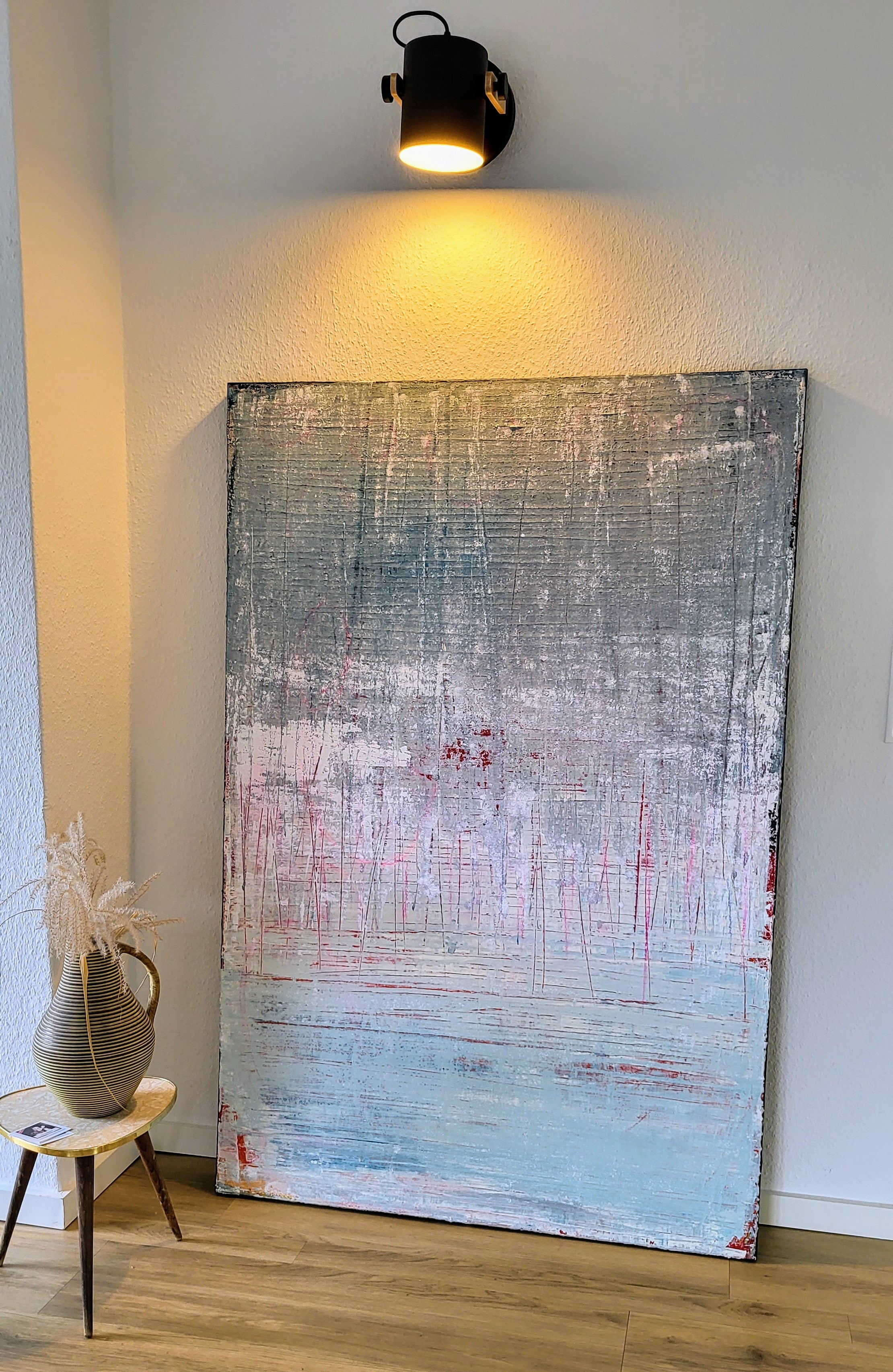 Vorschiedene Weiten von Conny Wachsmann, Künstlerin - ein großes abstraktes Bild in Pastellblau, Rosa und Weiß - lehnt vor einer weißen Wand unter Schwarzlicht. Ein kleiner Hocker mit getrockneten Gräsern vervollständigt die galerieartige Auslage auf dem Holzboden.