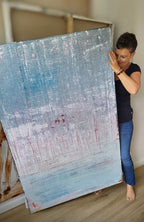 Eine Frau mit kurzen Haaren und Brille, barfuß, in schwarzem Hemd und blauer Jeans, hält "Vorschiedene Weiten", ein strukturiertes abstraktes Kunstwerk von Conny Wachsmann. Es wurde in Kratztechnik für die artvergnuegen galerie geschaffen und zeigt Blau-Weiß-Töne mit einem Hauch von Rot.