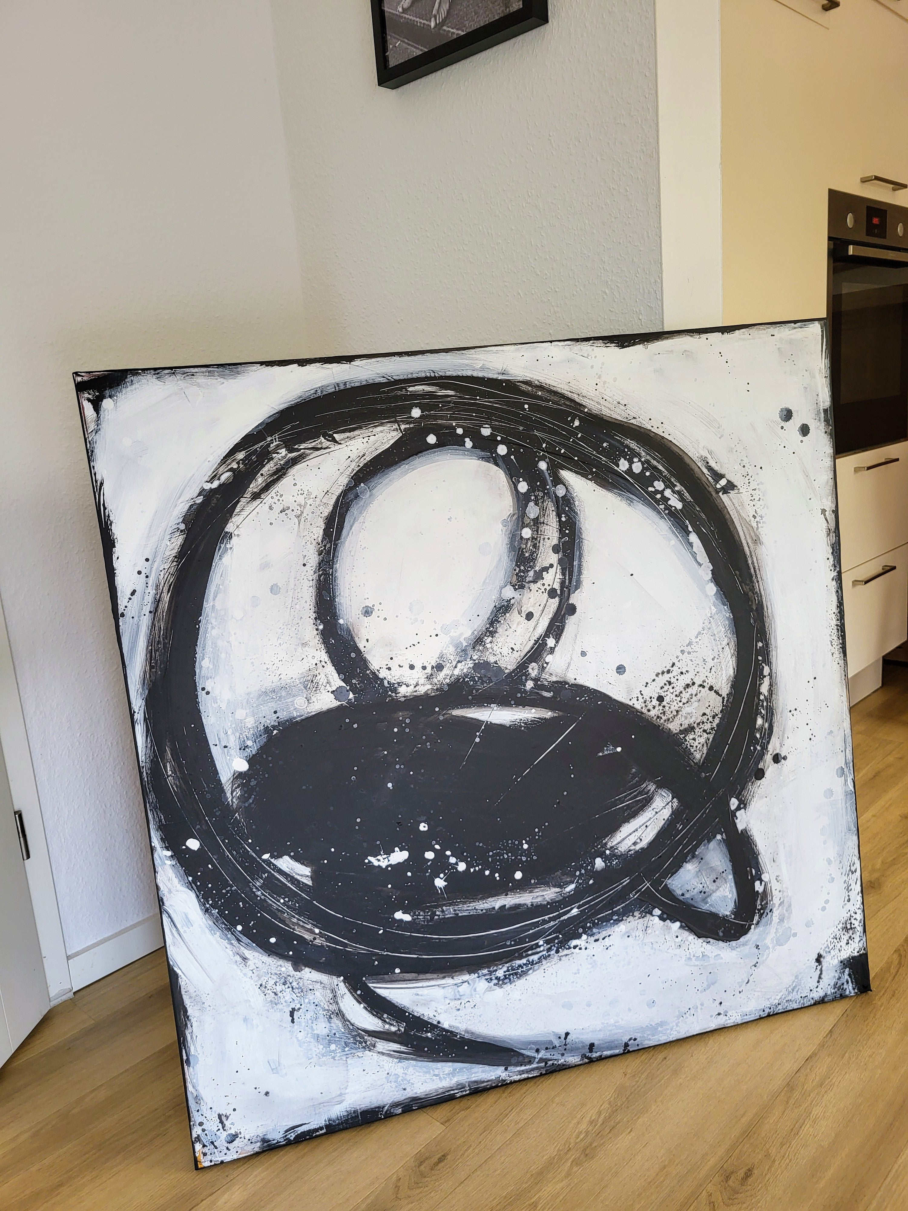 Nightswimming von Conny Wachsmann, Künstlerin, lehnt an einer Wand in einer modernen Küche. Kräftige, wirbelnde schwarze Pinselstriche, grobe Kreise und Farbspritzer heben sich von dem weißen Hintergrund ab.