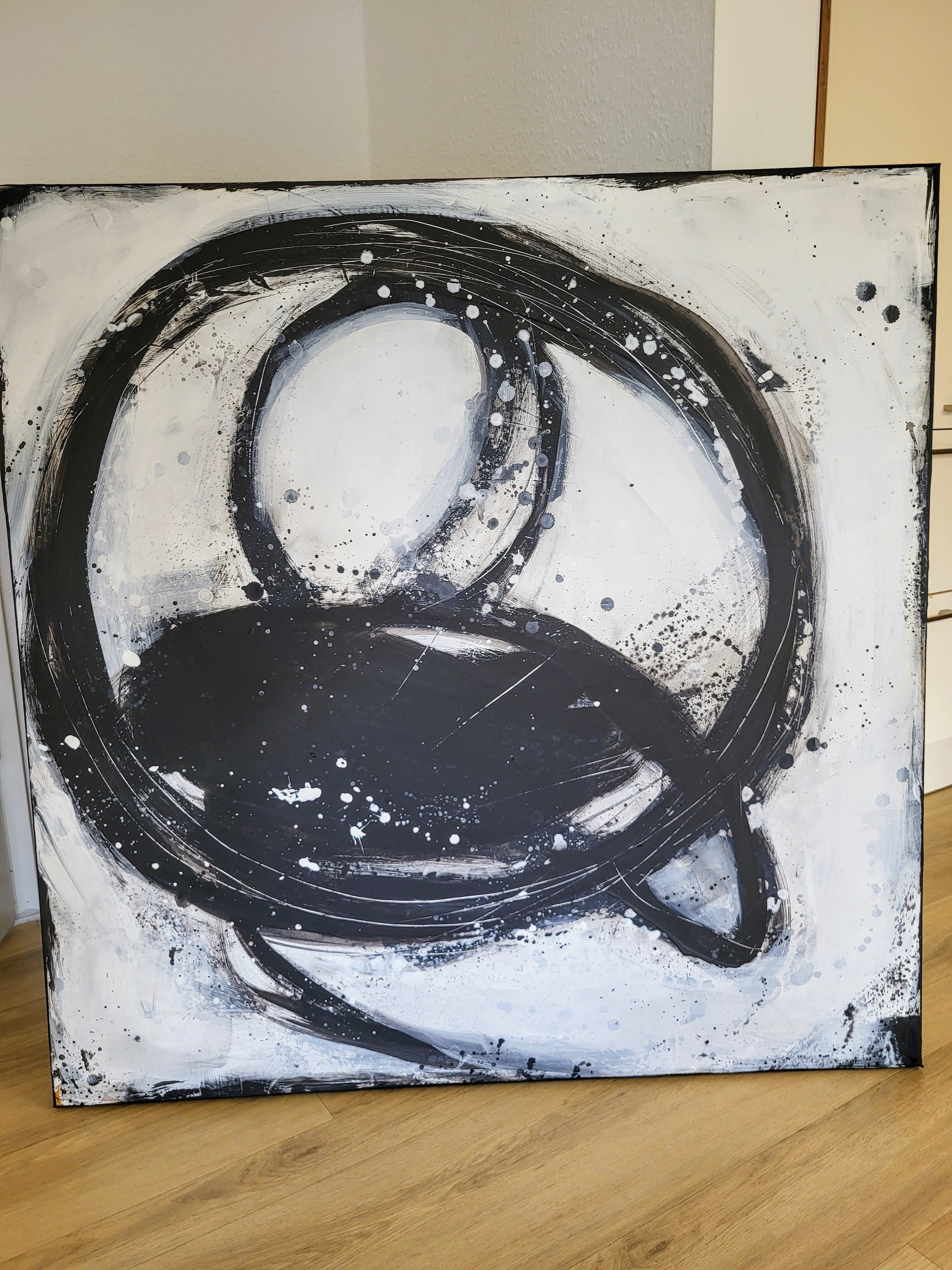 Nightswimming" von Conny Wachsmann, Künstlerin - ein auffälliges abstraktes Schwarz-Weiß-Gemälde mit kühnen, wirbelnden Kreisen und Spritzern auf einer quadratischen Leinwand - hebt sich von einer hellen Wand und einem Holzboden ab.