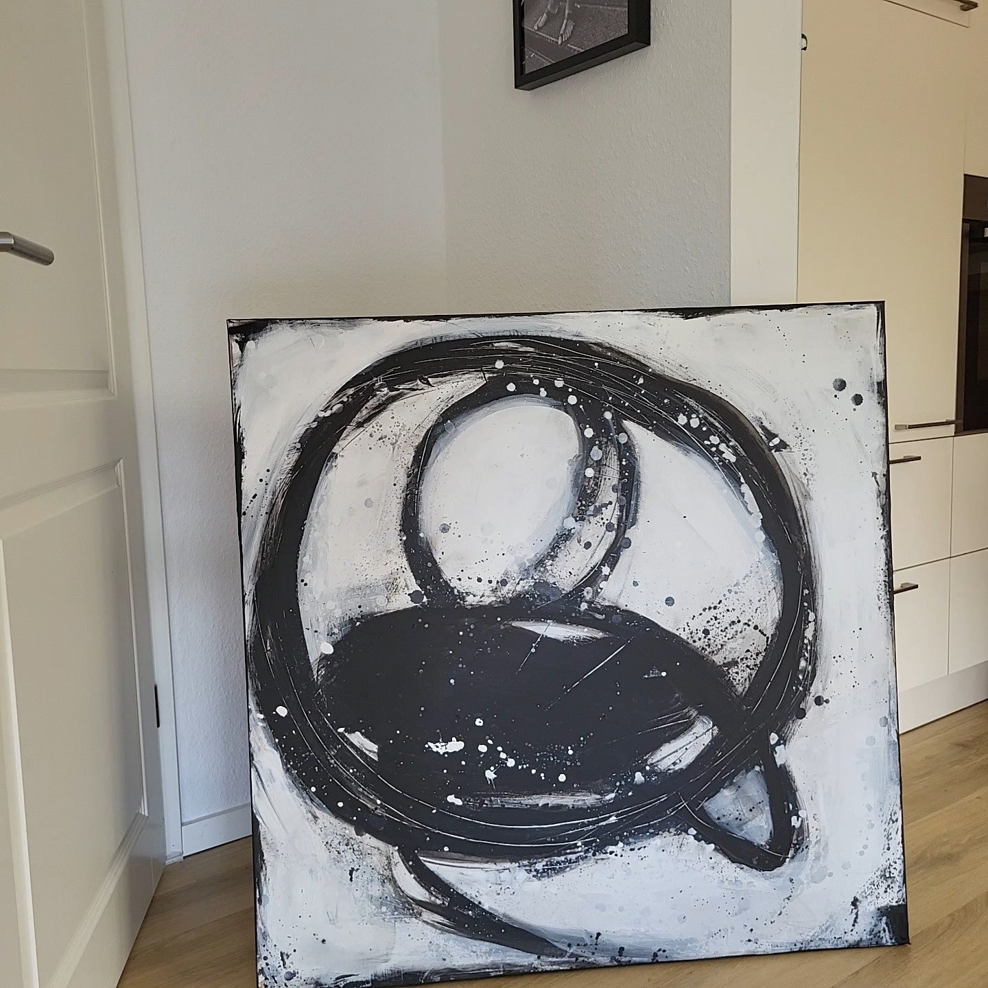 Nightswimming" von Conny Wachsmann, Künstlerin - ein abstraktes Kunstwerk mit kühnen schwarzen kreisförmigen und ovalen Pinselstrichen auf Weiß - lehnt an einer Wand in einem modernen Raum mit hellem Holzboden und weißen Wänden.