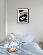 Eine flauschige weiße Katze sitzt auf einem ordentlich gemachten Bett mit hellblauen Laken; auf der Decke liegt ein Gebäckstück, und an der hellgrauen Wand über dem Bett hängt "Zeichnungen & Reduktion II" von Conny Wachsmann, Künstlerin.