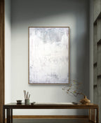 Ein minimalistisches Interieur zeigt "Stiller Spaziergang" von Conny Wachsmann, ein großes abstraktes Gemälde in sanftem Weiß und Grau über einem Holztisch mit Keramik, Stäbchen und einer Vase mit einem zarten Zweig für Ruhe und Entspannung.