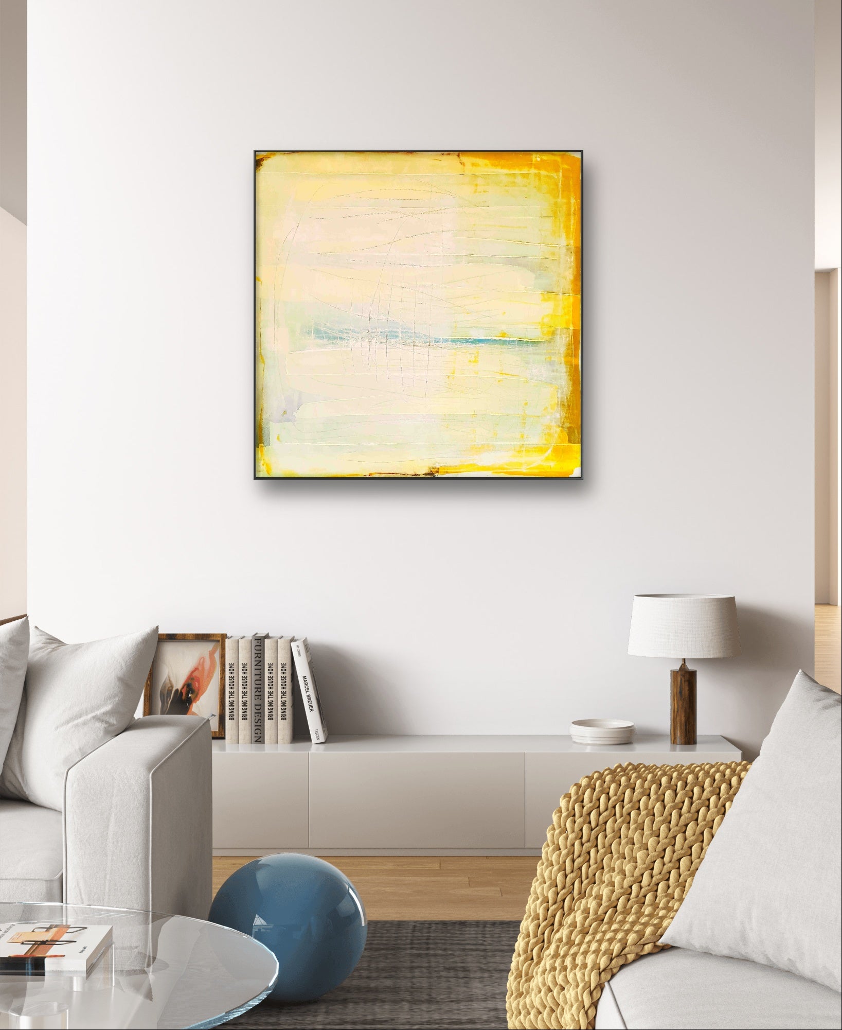 Ein modernes Wohnzimmer ist mit einem hellgrauen Sofa, einer Strickdecke, einer weißen Lampe und einem Bücherregal ausgestattet. Darüber hängt „Küstenzauber II“ von der Künstlerin Conny Wachsmann – eine große abstrakte Landschaft in Gelb und Weiß, die die weiße Wand aufhellt.