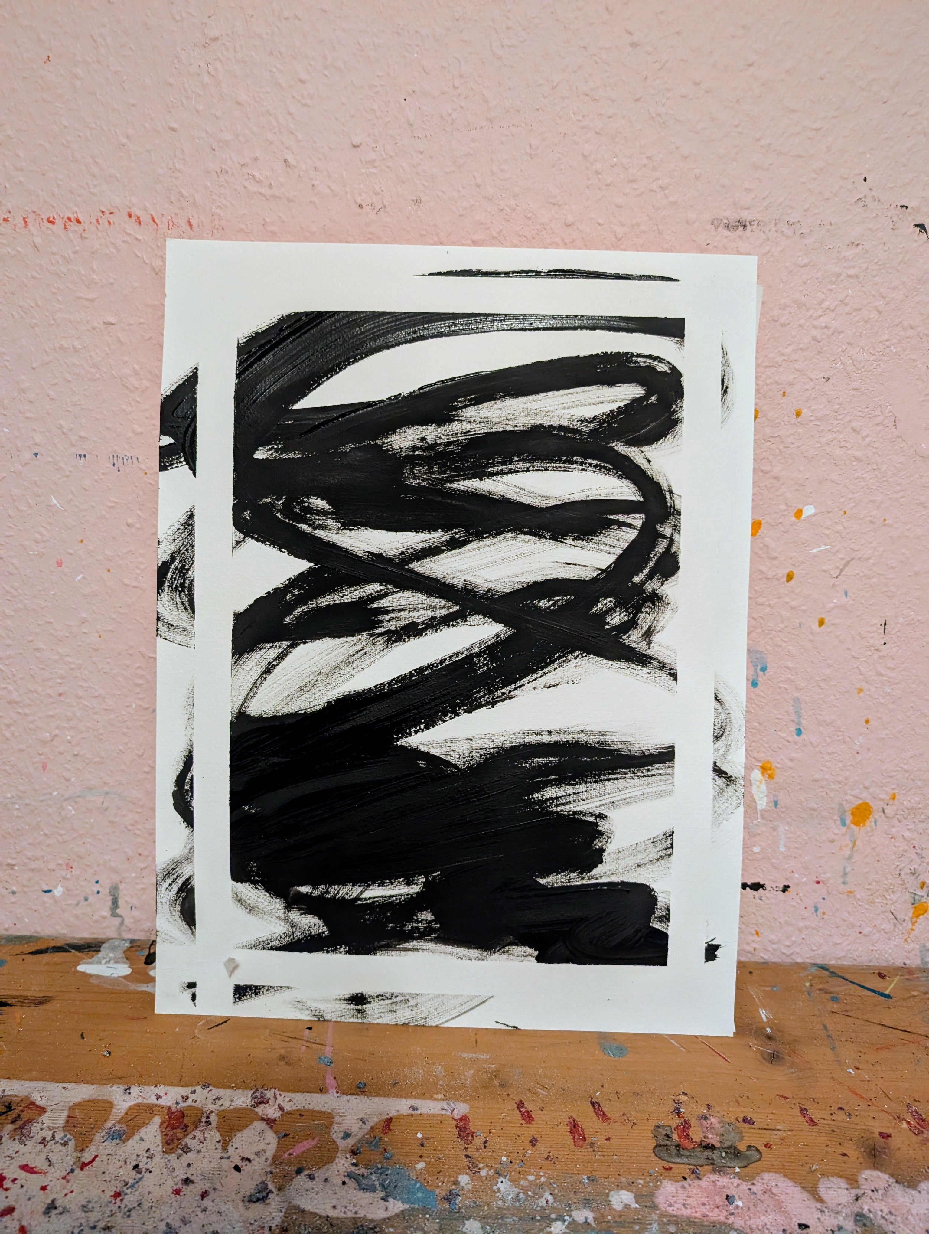 Zeichnungen & Reduktion III von Conny Wachsmann, Künstlerin - ein Blatt mit kühnen schwarzen abstrakten Pinselstrichen auf Weiß - ruht vor einer rosafarbenen Wand auf einer mit Farbe bespritzten Holzoberfläche und verkörpert minimalistische Kunst.
