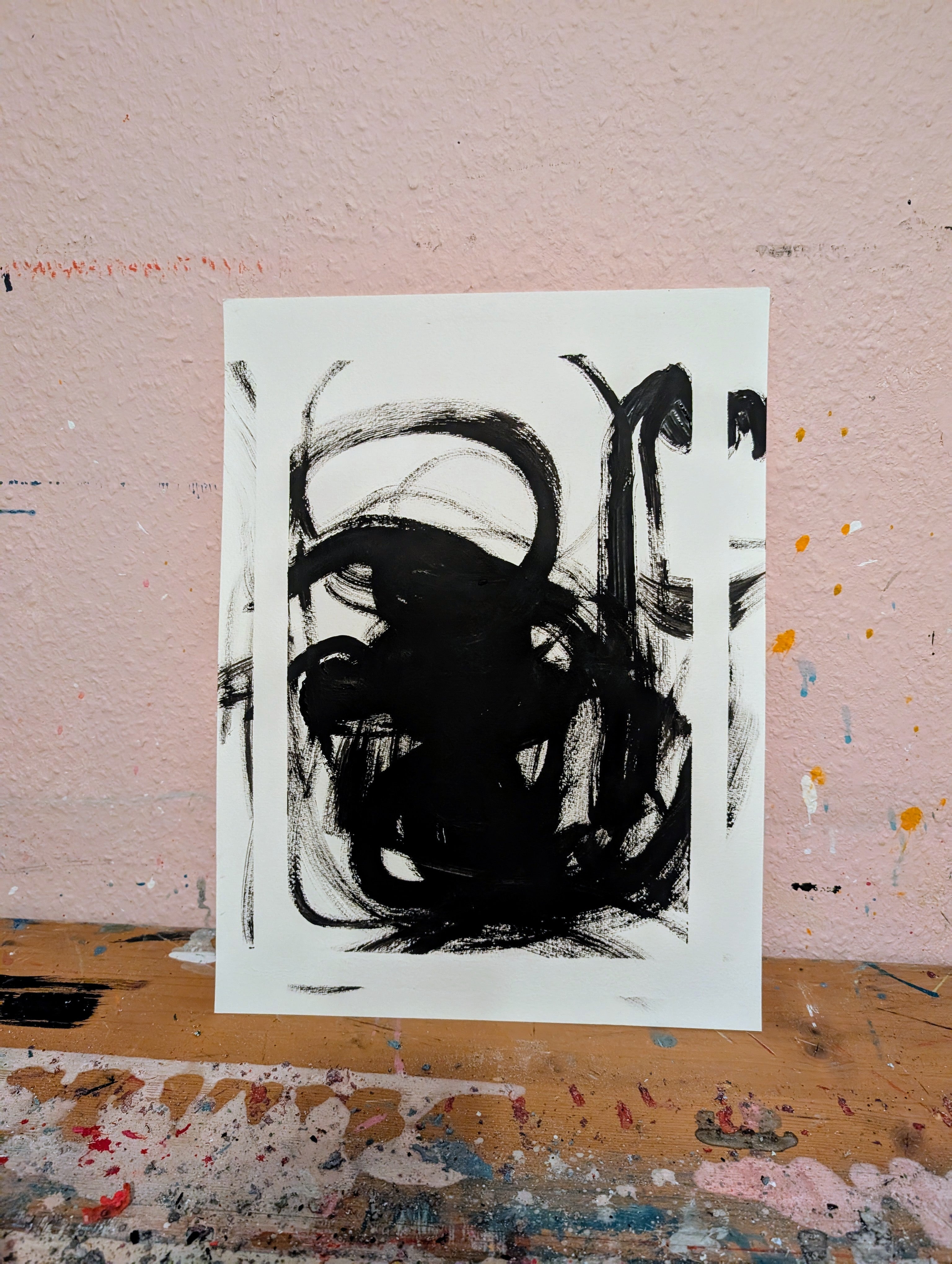 Zeichnungen & Reduktion IV" von Conny Wachsmann, Künstlerin: Ein weißes Papier mit kräftigen schwarzen abstrakten Pinselstrichen steht vor einer strukturierten hellrosa Wand auf einem mit Farbe bespritzten Holztisch.