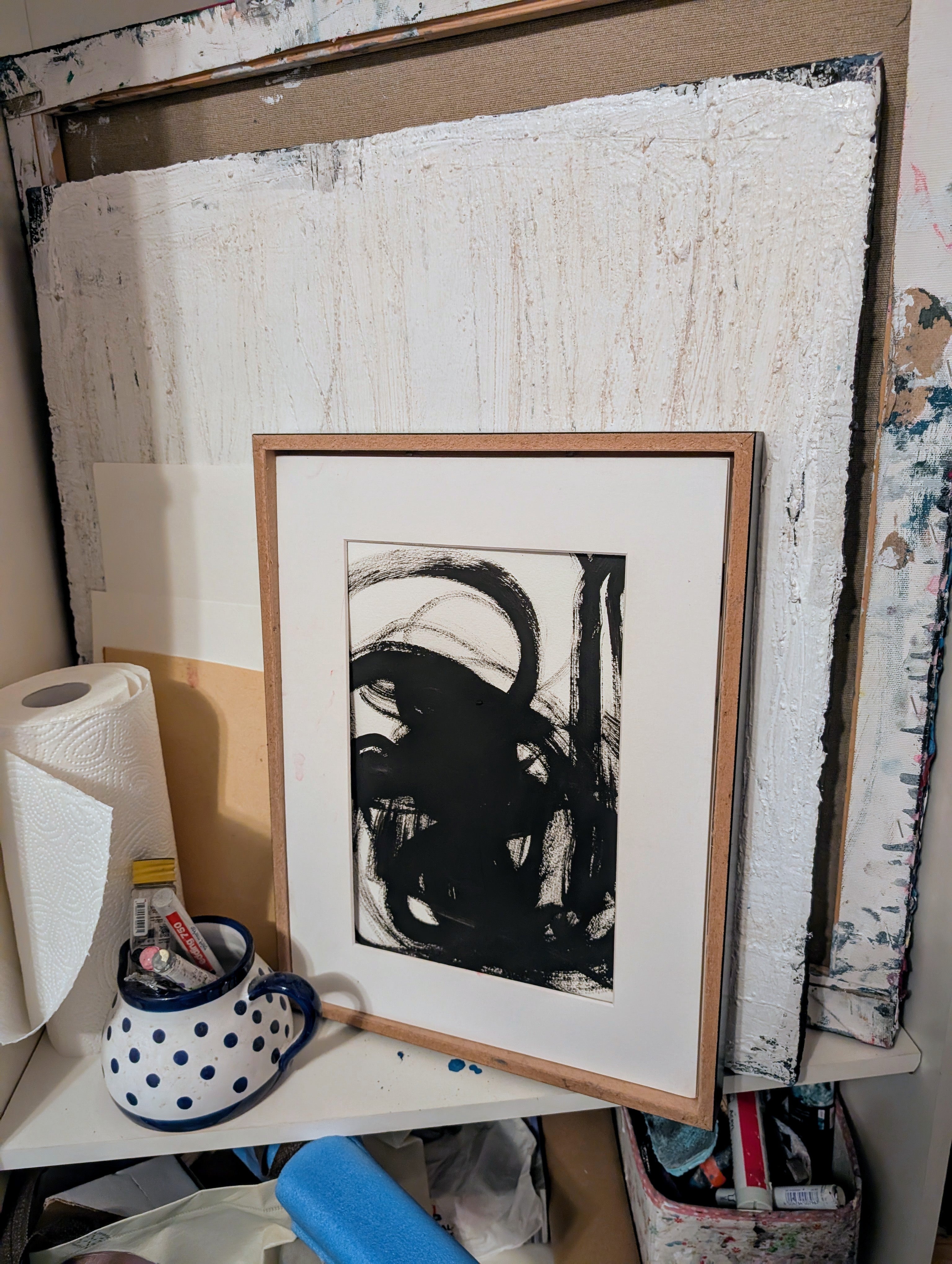 Die gerahmte Zeichnung "Zeichnungen & Reduktion IV" von Conny Wachsmann, Künstlerin, lehnt an weißen Leinwänden. Daneben stehen ein gepunkteter Becher mit Kunstmaterialien, eine Papierrolle und andere Utensilien auf einem Regal.