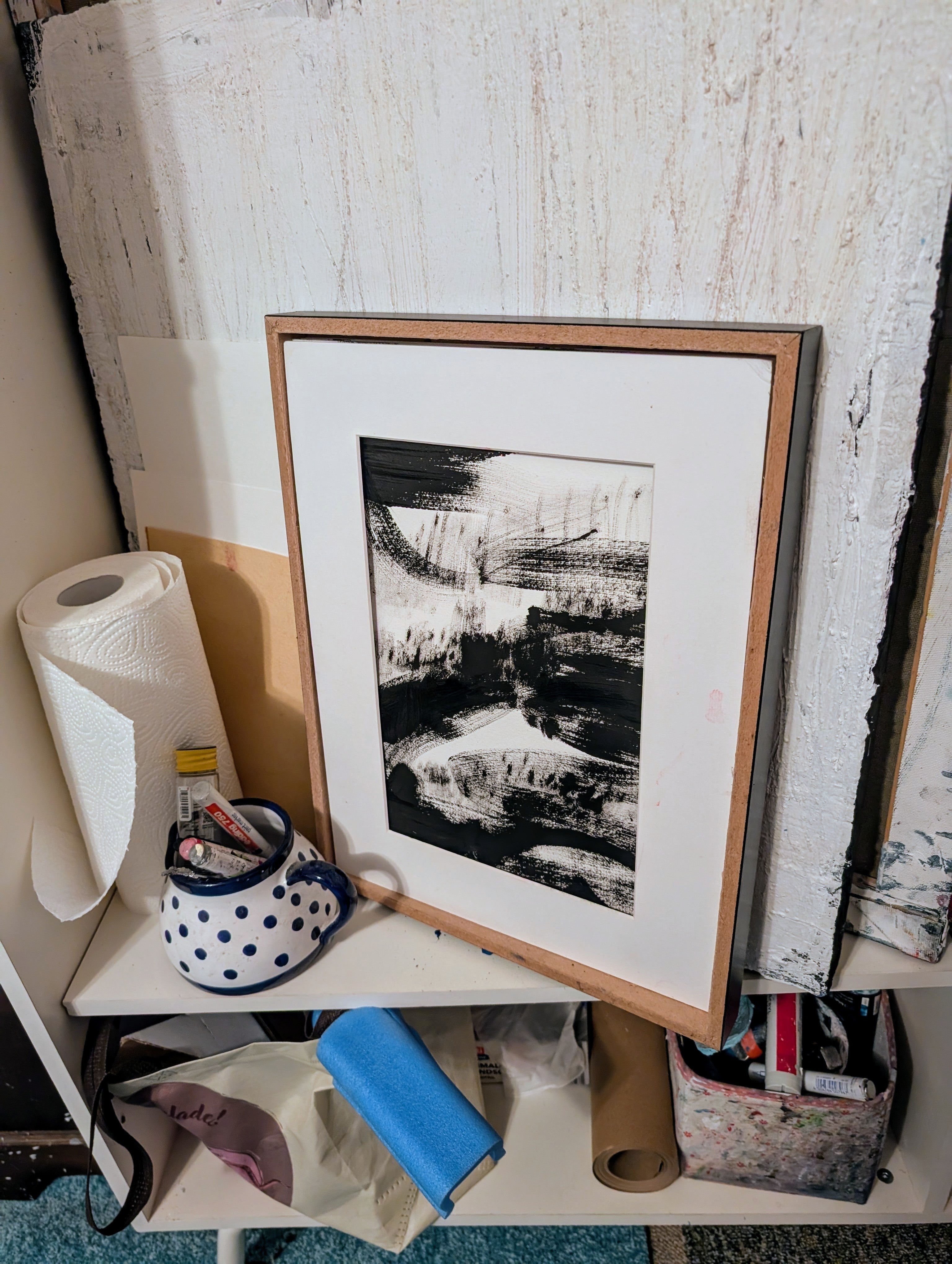 Das Kunstwerk "Zeichnungen & Reduktion II" von Conny Wachsmann, Künstlerin, ist gerahmt und lehnt an einer strukturierten Wand in einem Regal neben einer Rolle Papierhandtücher, einem blauen Tuch, einem gepunkteten Becher mit Pinseln und diversem Kunstzubehör.