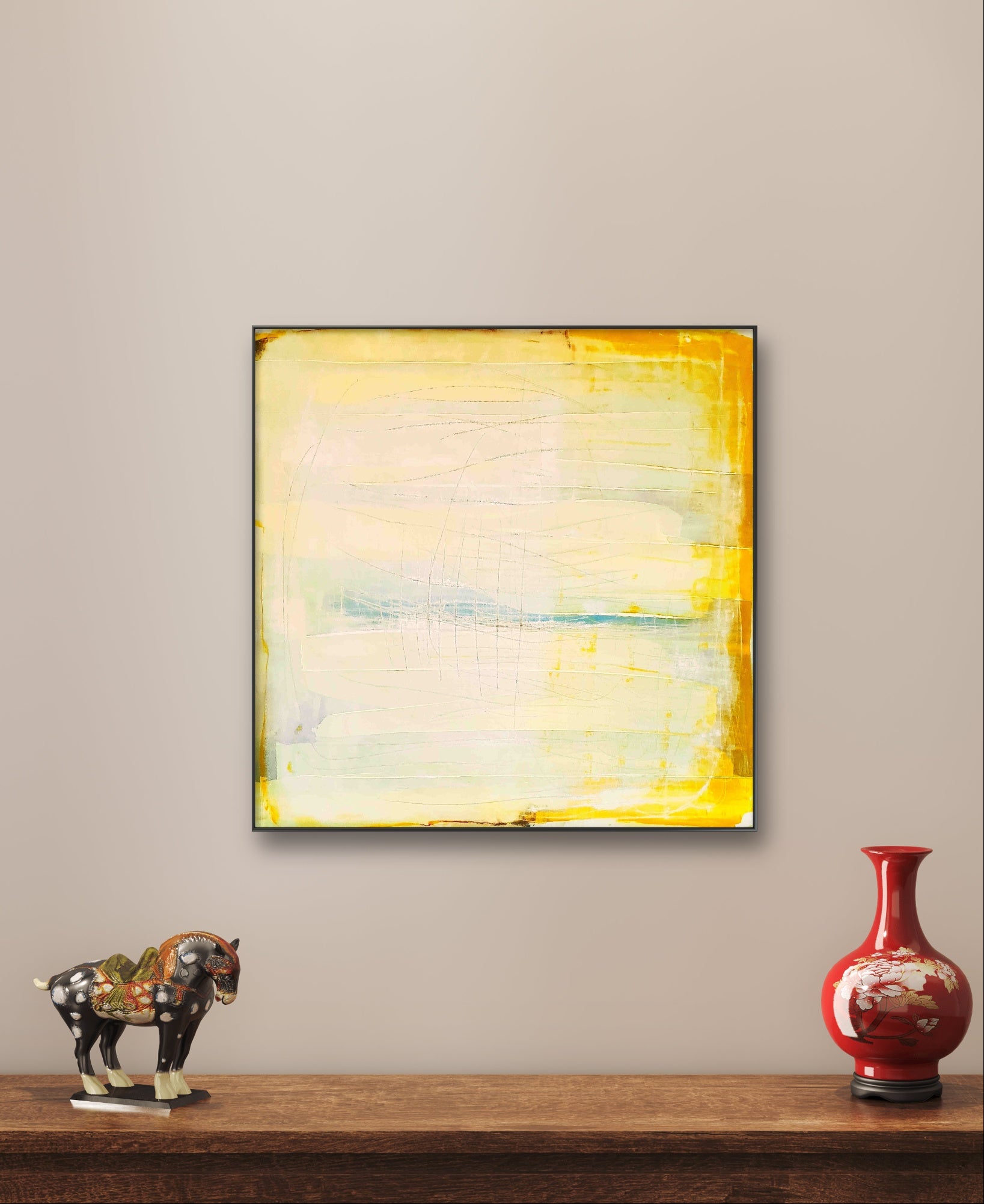 Küstenzauber II, ein quadratisches abstraktes Landschaftskunstwerk der Künstlerin Conny Wachsmann mit gelben Rändern, hängt an einer beigen Wand über einem Holzregal mit einem Keramikpferd und einer roten Vase mit weißem Blumenmuster.