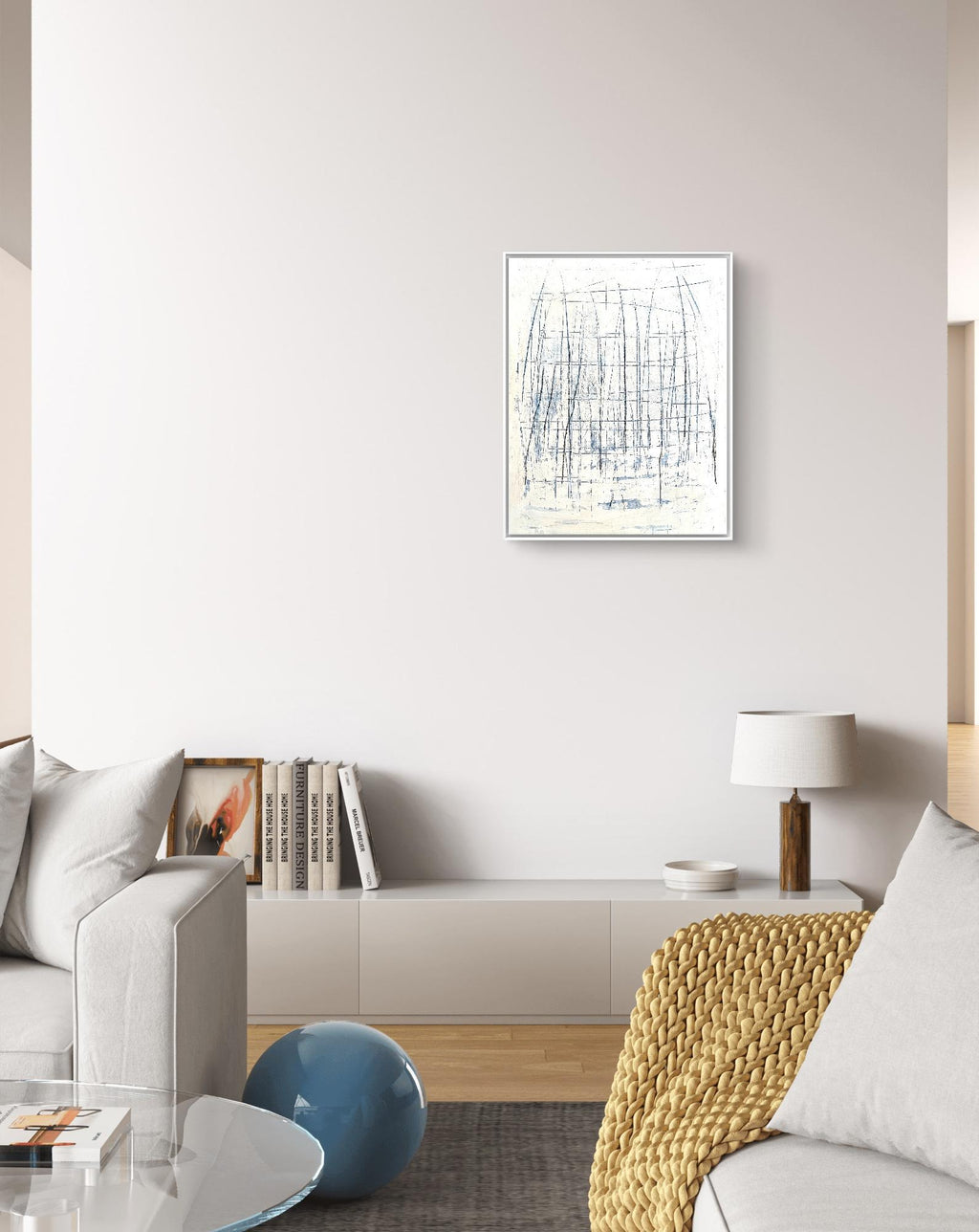 Ein modernes Wohnzimmer mit hellgrauen Sofas, einem niedrigen weißen Schrank, Büchern, einer Tischlampe, einem Couchtisch aus Glas, einer blauen Dekokugel und „Feldlinien“ von Conny Wachsmann, Künstlerin, als abstrakte Kunst an der weißen Wand ausgestellt.