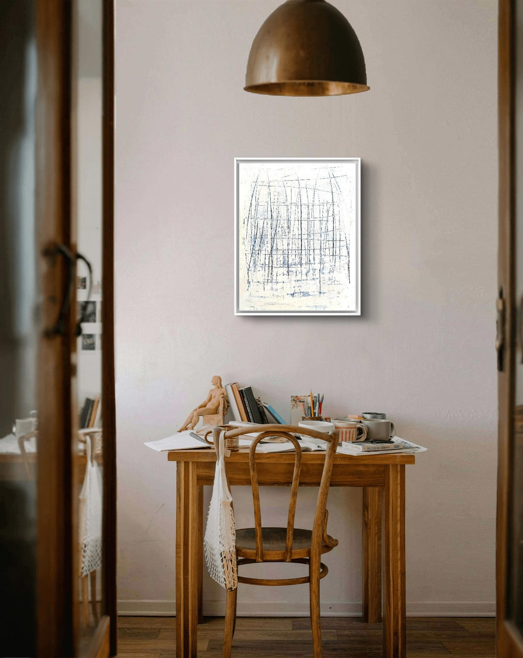 Ein gemütlicher Holzschreibtisch mit Künstlerutensilien, einer Tasse und einer Schaufensterpuppe steht an einer hellen Wand. Darüber hängt „Feldlinien“, ein Acrylbild auf Leinwand der Künstlerin Conny Wachsmann, ergänzt durch eine braune Hängelampe und einen Holzstuhl.