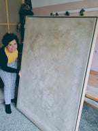 Eine Person mit lockigem Haar, in einem gelben Oberteil und einer karierten Hose, lächelt im Innenbereich in die Kamera und hält dabei in beiden Händen „Spurensuche“ der Künstlerin Conny Wachsmann.