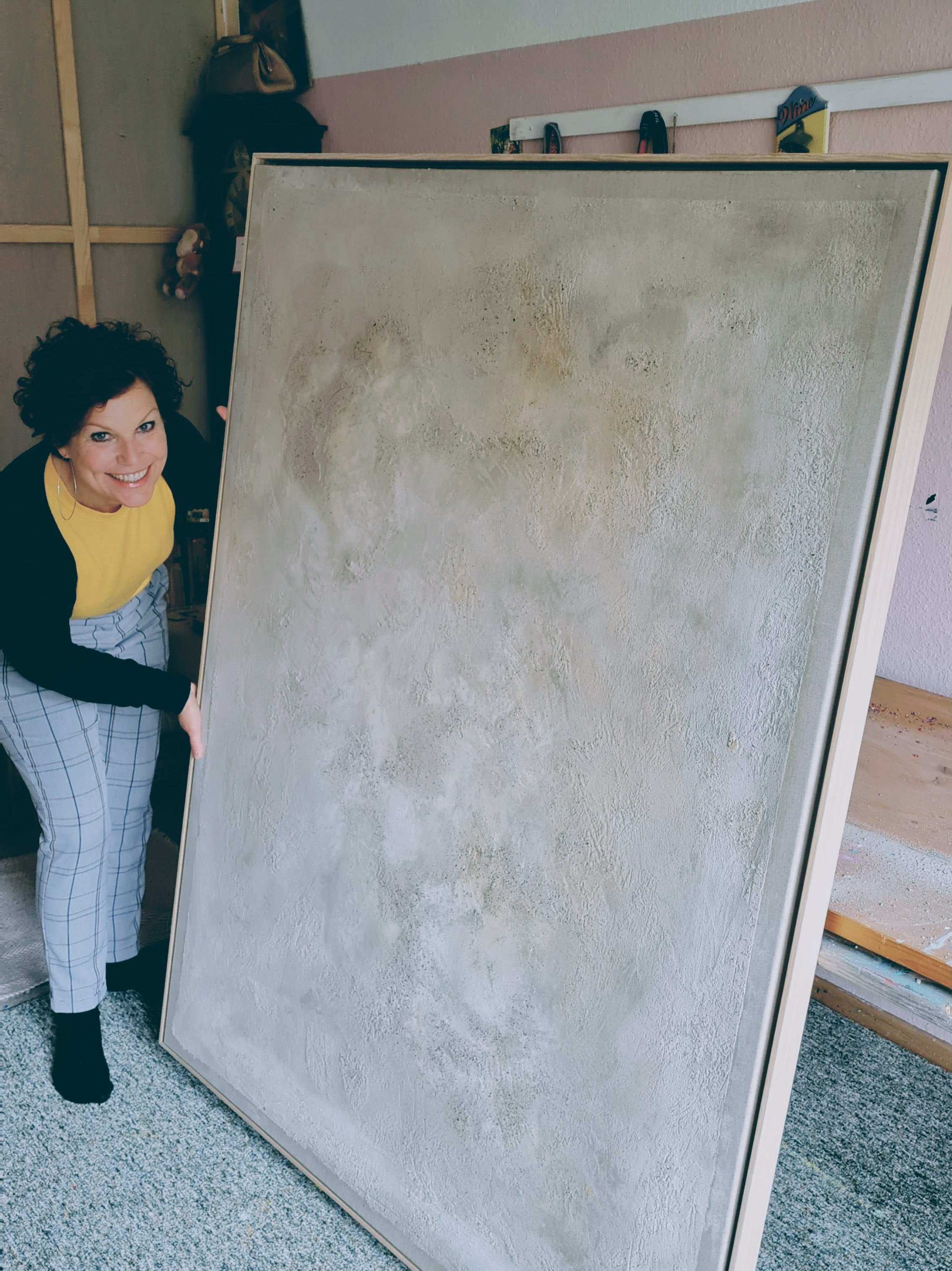 Eine Person mit lockigem Haar, in einem gelben Oberteil und einer karierten Hose, lächelt im Innenbereich in die Kamera und hält dabei in beiden Händen „Spurensuche“ der Künstlerin Conny Wachsmann.