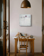 Ein Holzschreibtisch mit Künstlerutensilien und einer Tasse steht an einer hellen Wand, über der „Stille Spur“ von Conny Wachsmann hängt. Ein Holzstuhl und eine Kupferpendelleuchte runden diesen gemütlichen, künstlerischen Raum ab.