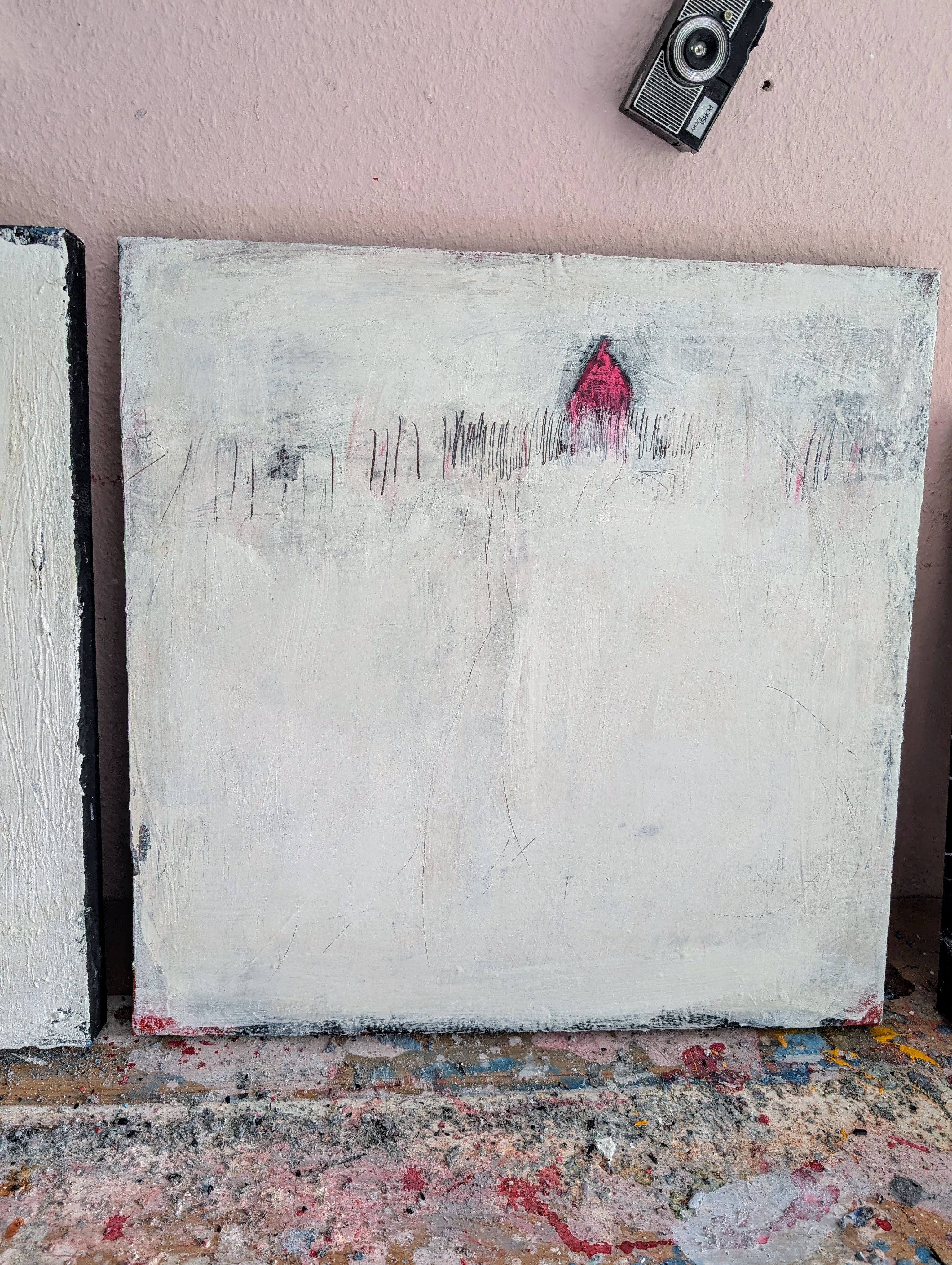 „Stille Spur“ von der Künstlerin Conny Wachsmann ist ein überwiegend weißes abstraktes Originalgemälde mit schwachen Linien, Flecken und einem kleinen Haus mit rotem Dach im oberen Bereich. Das Unikat lehnt an einer rosa Wand unter einer Vintage-Kamera.