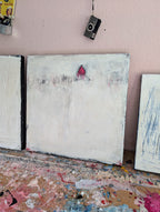 „Stille Spur“, ein großes, größtenteils weißes abstraktes Originalgemälde von Conny Wachsmann, zeigt in der Mitte ein kleines rotes Haus und lehnt an einem farbbespritzten Boden neben einzigartigen Leinwänden und einer Vintage-Kamera an einer hellrosa Wand.