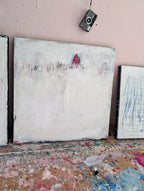 Das minimalistische Unikat „Stille Spur“ von Conny Wachsmann, Künstlerin, zeigt ein rotes Haus in verschneiter Landschaft vor einer Wand; Farbspritzer bedecken den Arbeitsplatz, darüber hängt eine Vintage-Kamera.