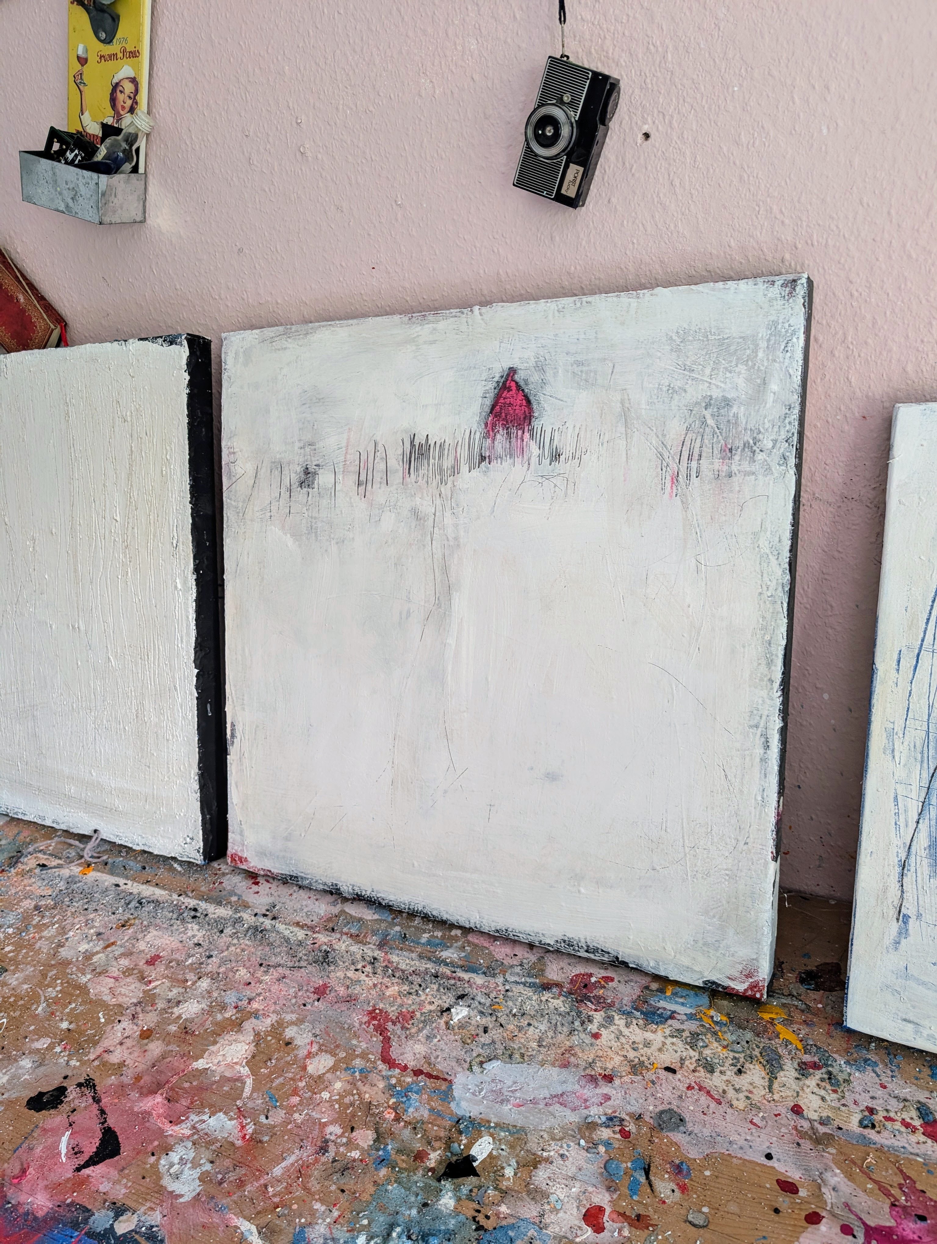 „Stille Spur“ von Conny Wachsmann zeigt eine verschneite Landschaft mit einem roten Haus vor einer farbbesprenkelten Fläche, darüber an der blassrosa Wand hängen eine alte Kamera und andere Gegenstände.