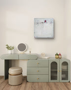 Ein minimalistischer Schminktisch mit einem salbeigrünen Schreibtisch, einem runden Spiegel, einem Hocker und einer kleinen Pflanze. Darüber hängt „Stille Spur“ von Conny Wachsmann – ein originales strukturiertes weiß-rotes Gemälde – an der beigefarbenen Wand. Holzboden rundet das moderne Dekor ab.