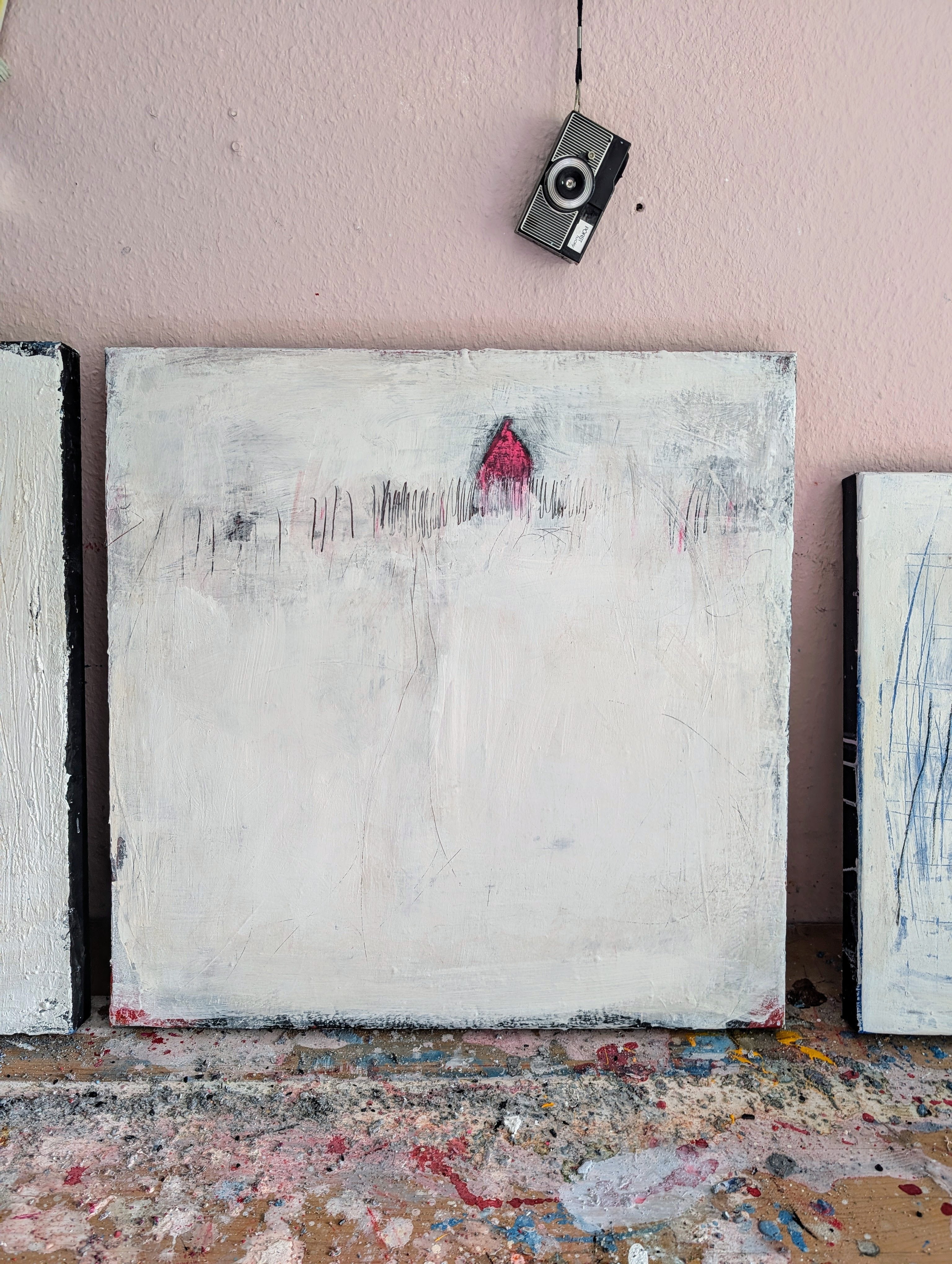 „Stille Spur“ von Conny Wachsmann: Ein minimalistisches Originalgemälde mit weißem Hintergrund, feinen Linien und einem kleinen roten Haus. Vintage-Kamera und rosafarbener Wand, Farbspritzer bedecken die Fläche darunter.