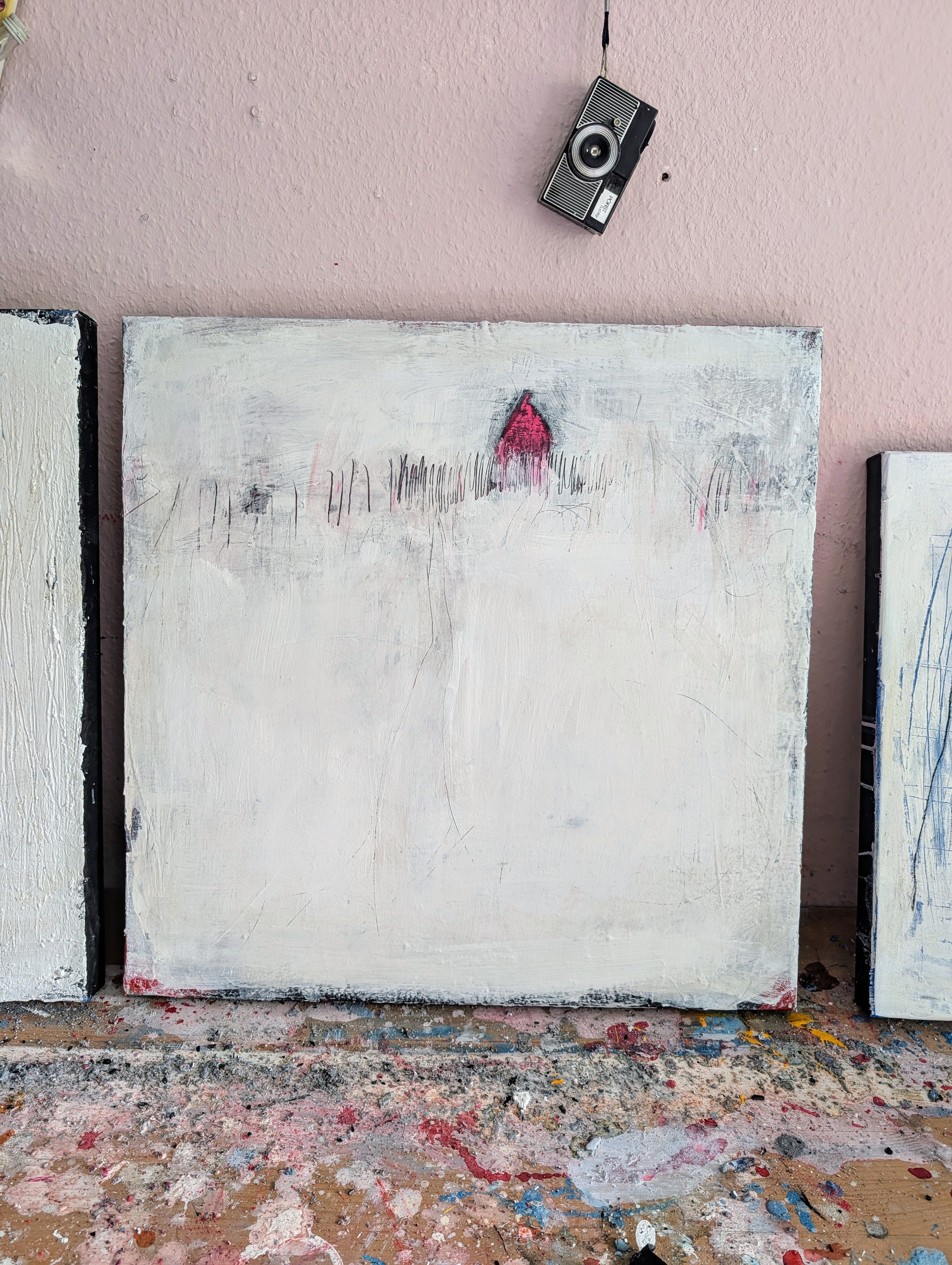 „Stille Spur“ von Conny Wachsmann ist ein minimalistisches abstraktes Originalgemälde mit überwiegend weißem Hintergrund, dezenten grauen Flecken und einem kleinen roten Dreieck oben in der Mitte. Darüber hängt eine alte Kamera an einer rosa Wand; Farbspritzer zieren den Boden.