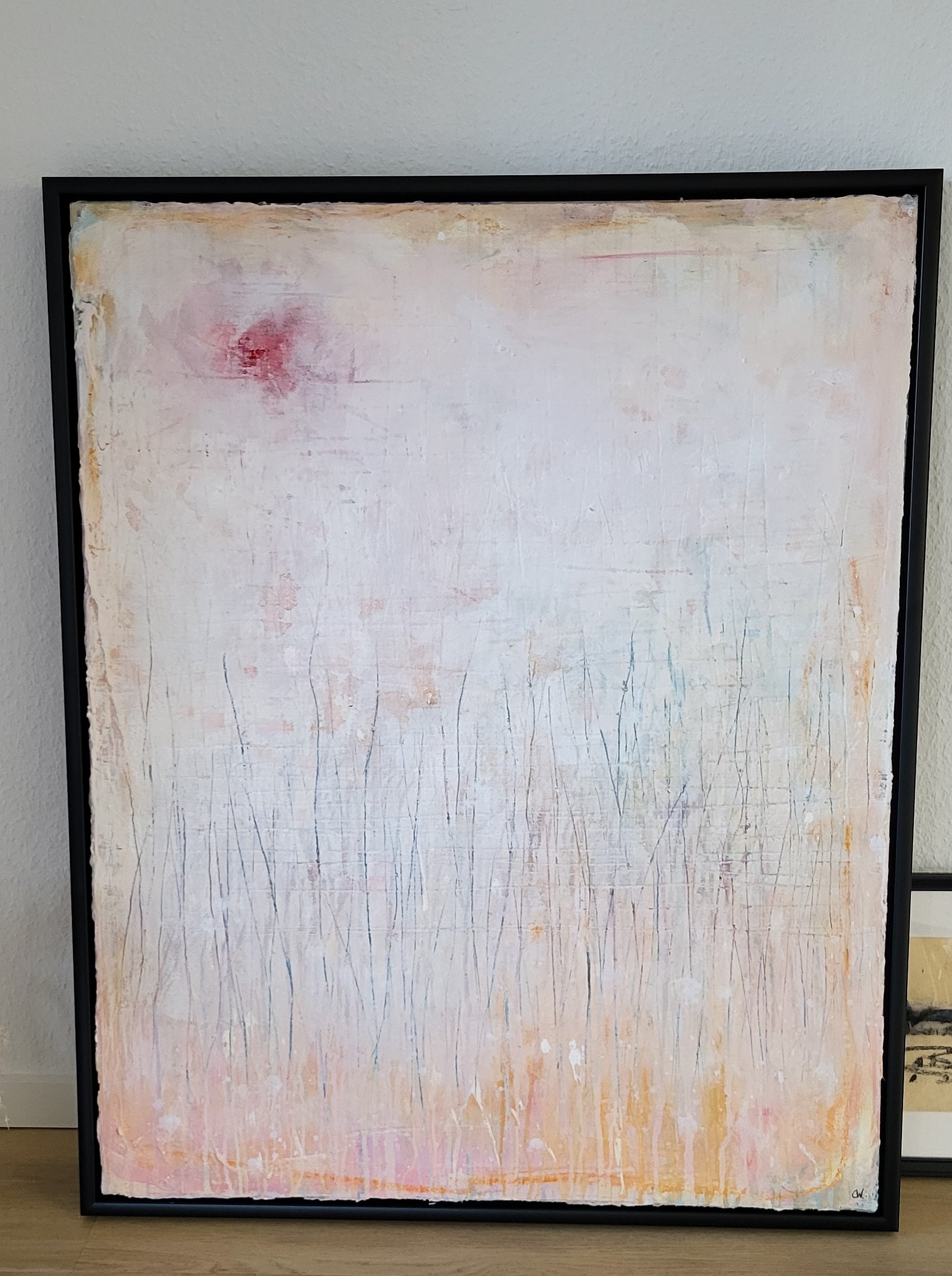 „Gedankenregen“ von Conny Wachsmann, ein einzigartiges gerahmtes abstraktes Gemälde in blassem Rosa und Weiß mit schwachen vertikalen Linien, sanften Orange- und Gelbtönen am unteren Rand und einem verschwommenen roten Fleck in der oberen linken Ecke, lehnt an einer Wand.