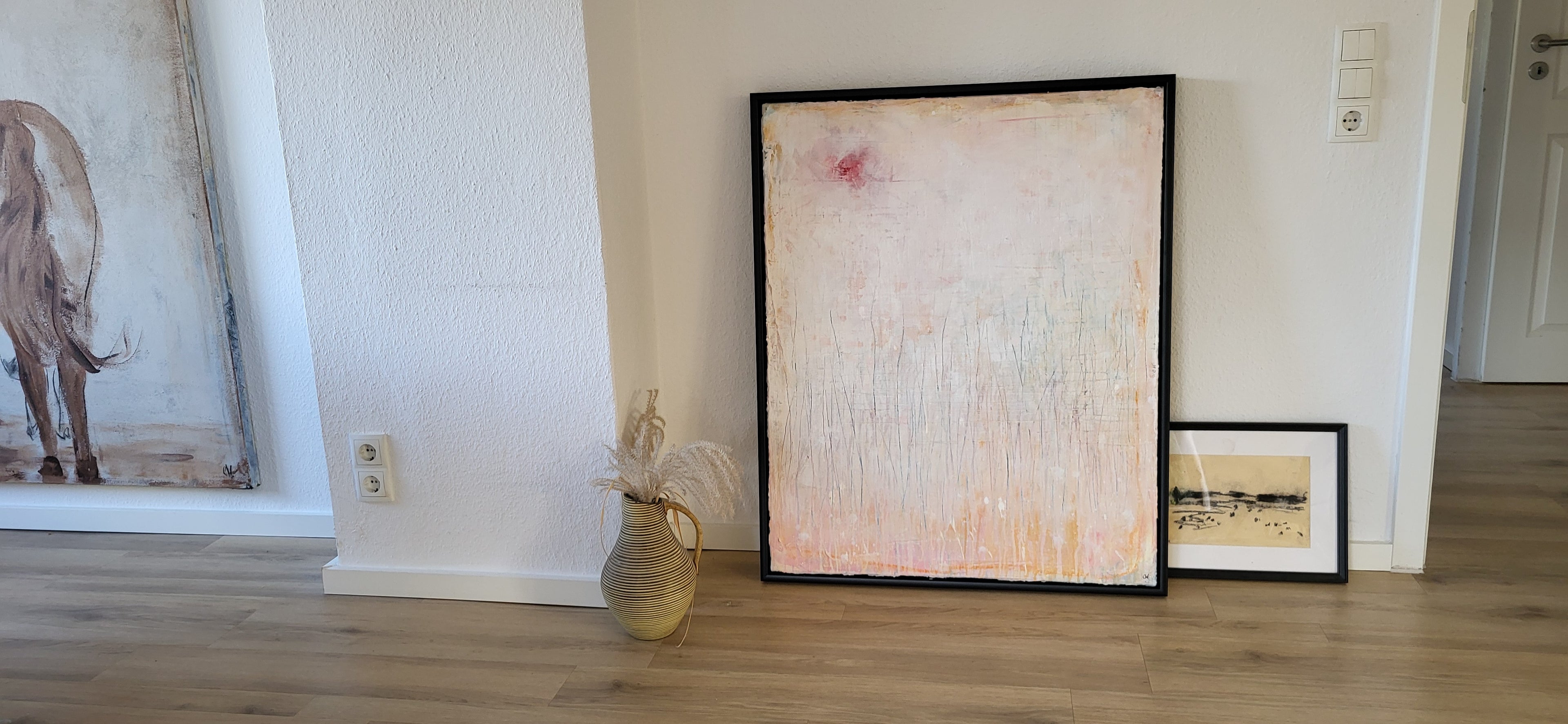 Das große Unikat „Gedankenregen“ von Conny Wachsmann in zarten Pastelltönen lehnt an einer weißen Wand neben einem kleineren gerahmten Bild, einer dekorativen Vase mit getrockneten Gräsern und links einem Teil eines Pferdegemäldes.