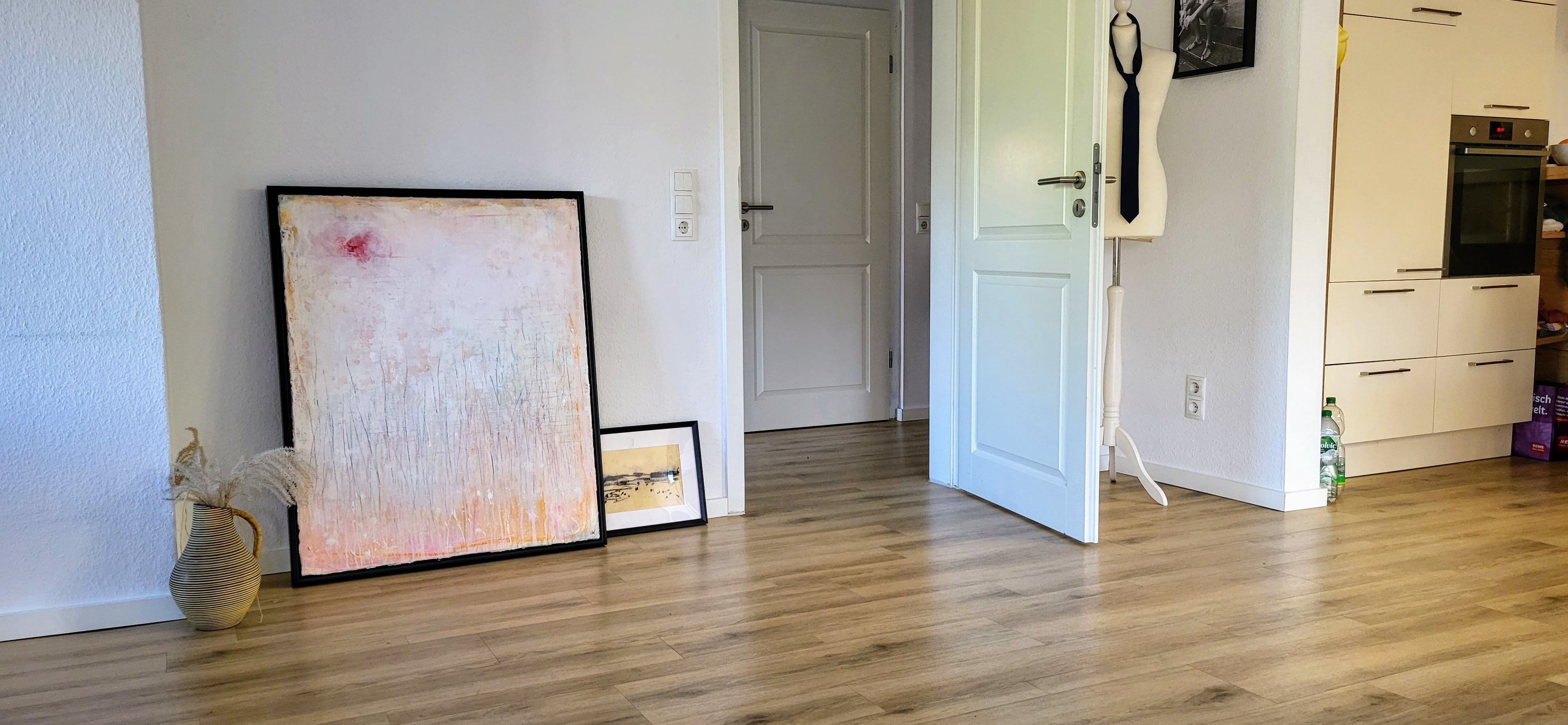Ein moderner Raum mit hellem Holzboden. Zwei einzigartige „Gedankenregen“-Auftragsgemälde von Conny Wachsmann lehnen gerahmt an der Wand neben einer Vase mit getrockneten Pflanzen. Eine offene Tür, eine Krawatte an der Garderobe und ein Blick in die Küche sind zu sehen.