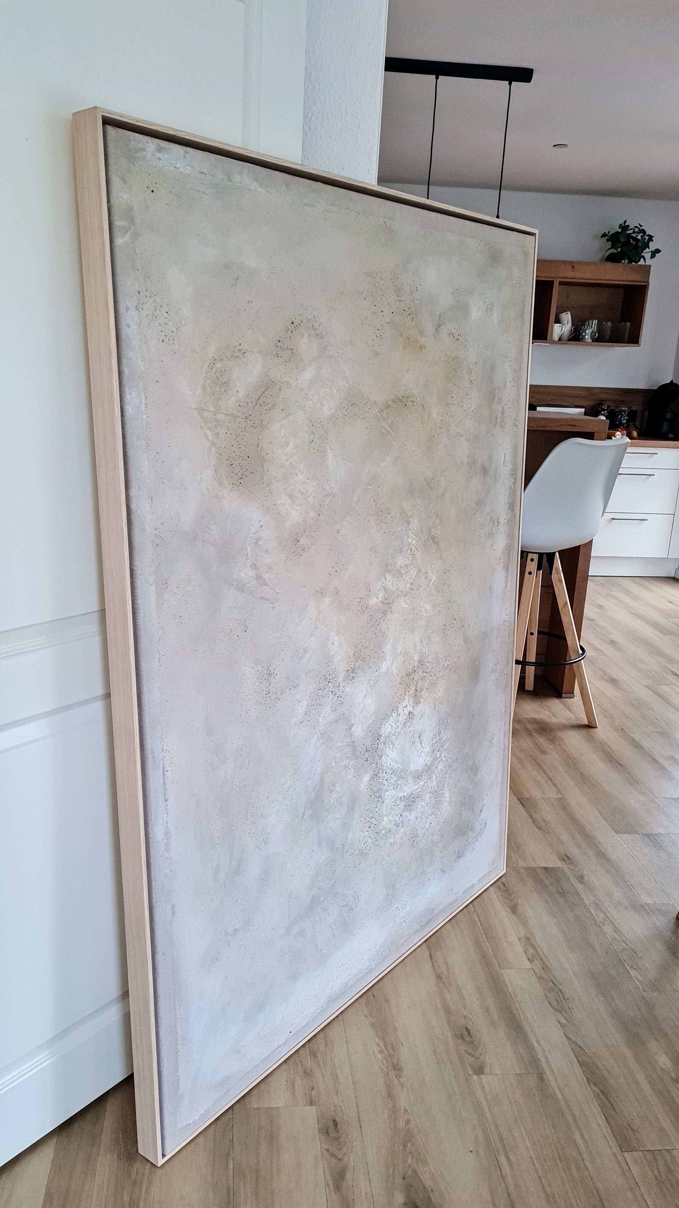 „Spurensuche“ von Conny Wachsmann, Künstlerin – ein großes abstraktes Kunstwerk in hellen, strukturierten Neutraltönen, eingerahmt in einen Schattenfugenrahmen – lehnt an einer weißen Wand auf einem Holzboden in einem modernen Küchen- und Essbereich.
