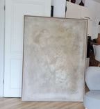 „Spurensuche“, ein großes gerahmtes abstraktes Kunstwerk von Conny Wachsmann, zeichnet sich durch strukturierte Beige- und Cremetöne aus. Es lehnt an einer weißen Tür in einem modernen Raum mit hellem Holzboden, in der Nähe eines grauen Stuhls und Schattenfugenrahmen-Schränken.