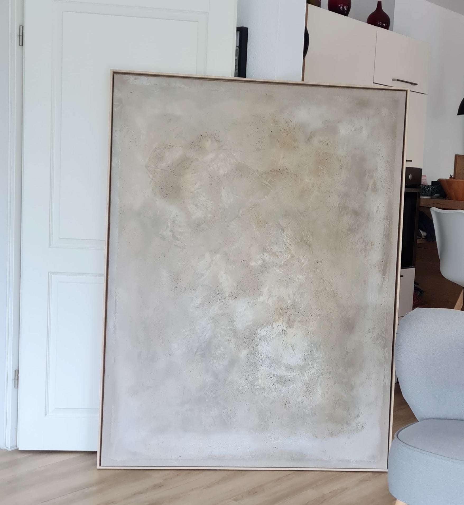 „Spurensuche“, ein großes gerahmtes abstraktes Kunstwerk von Conny Wachsmann, zeichnet sich durch strukturierte Beige- und Cremetöne aus. Es lehnt an einer weißen Tür in einem modernen Raum mit hellem Holzboden, in der Nähe eines grauen Stuhls und Schattenfugenrahmen-Schränken.
