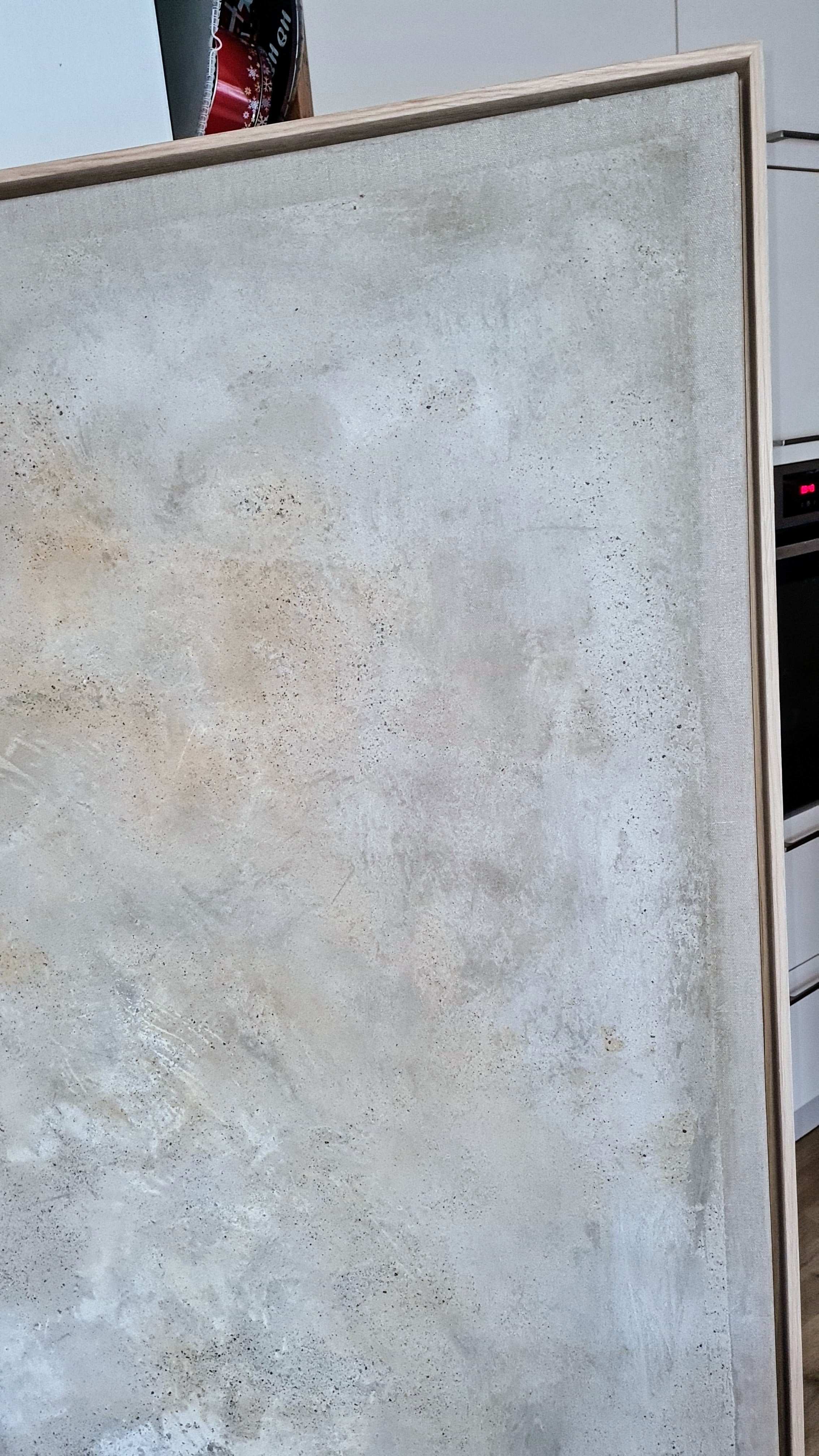 Das große gerahmte abstrakte Kunstwerk „Spurensuche“ von Conny Wachsmann in strukturierten Beige-, Grau- und Weißtönen lehnt an einer Küchentheke mit einem Backofen und roten Kanistern im Hintergrund.