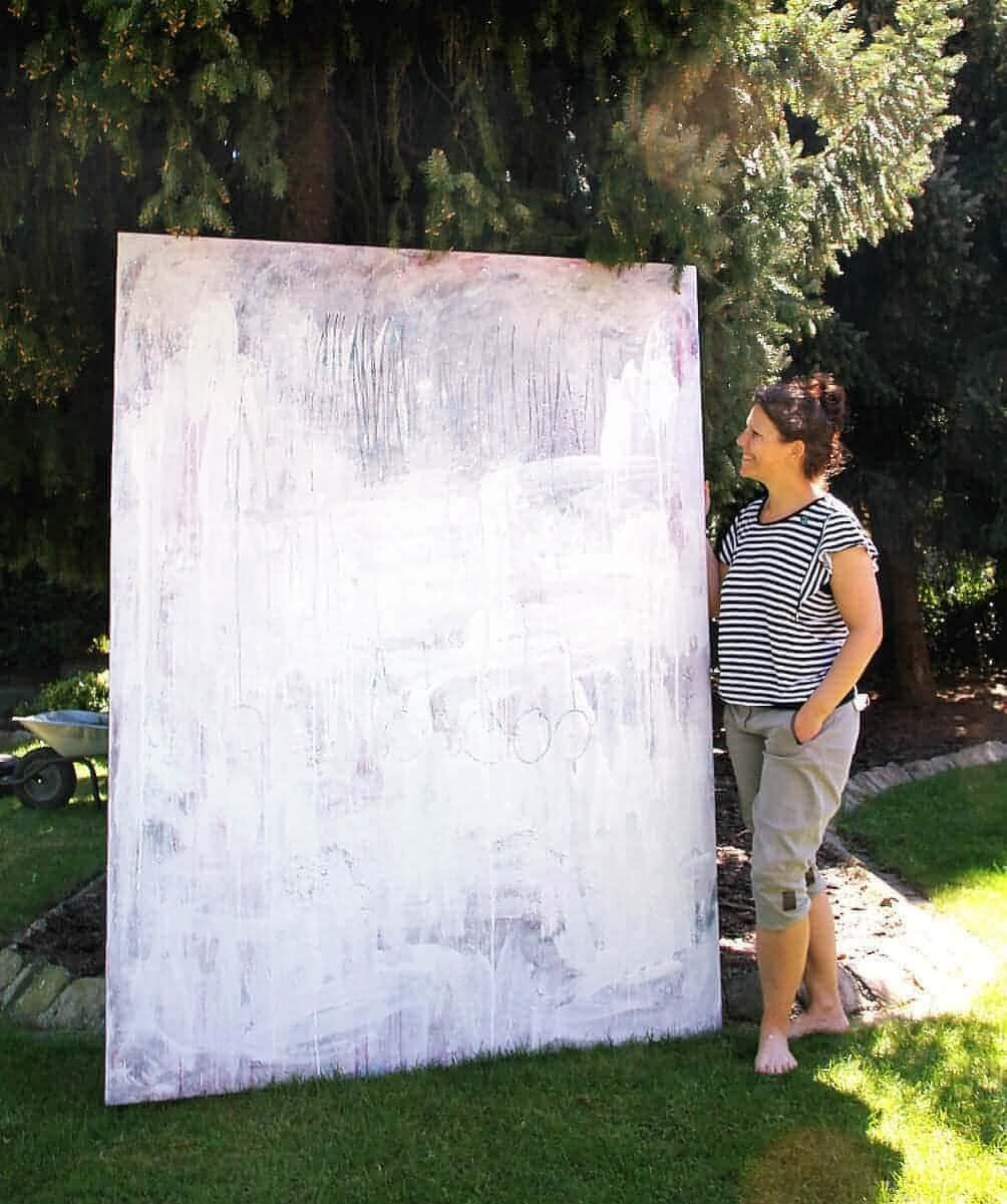 Eine Frau in gestreiftem Hemd und hochgekrempelten Hosen steht barfuß im Gras und lächelt auf "Stiller Spaziergang" von Conny Wachsmann - ein großes abstraktes Gemälde in weißen und hellgrauen Tönen, das im Freien für eine ruhige, entspannte Atmosphäre sorgt.