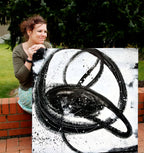 Eine Frau mit lockigem Haar lächelt, während sie "Nightswimming" von Conny Wachsmann, Künstlerin, in der Hand hält - ein großes abstraktes Gemälde mit kühnen schwarzen, wirbelnden Linien und Spritzern auf weißem Grund - während sie draußen auf einer Backsteinmauer mit Grün hinter ihr sitzt.