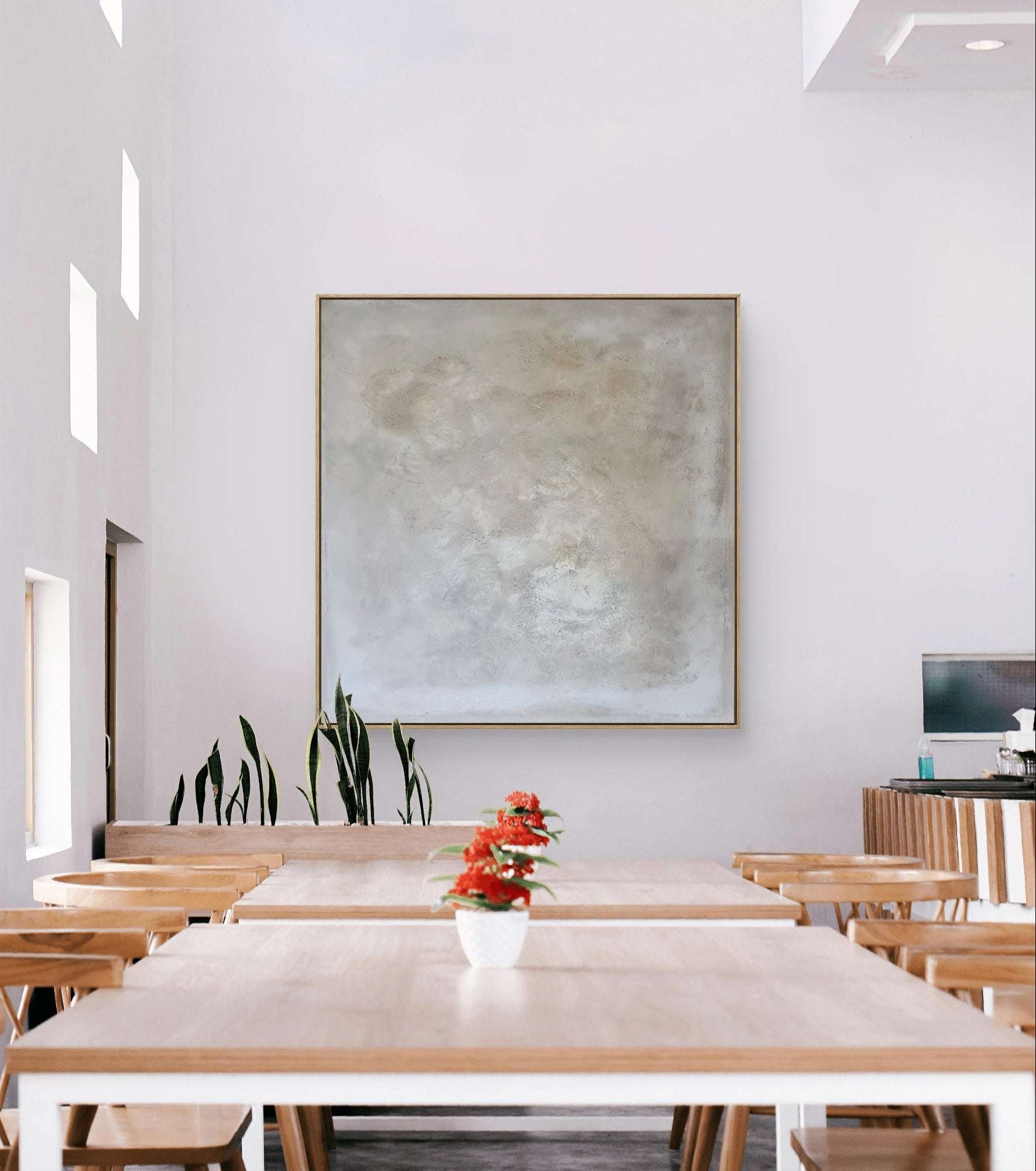 Heller, moderner Essbereich mit hellen Holztischen und Stühlen, roten Blumentöpfen, hohen grünen Pflanzen an der Wand und „Spurensuche“ von Conny Wachsmann in einem Schattenfugenrahmen an der weißen Wand.