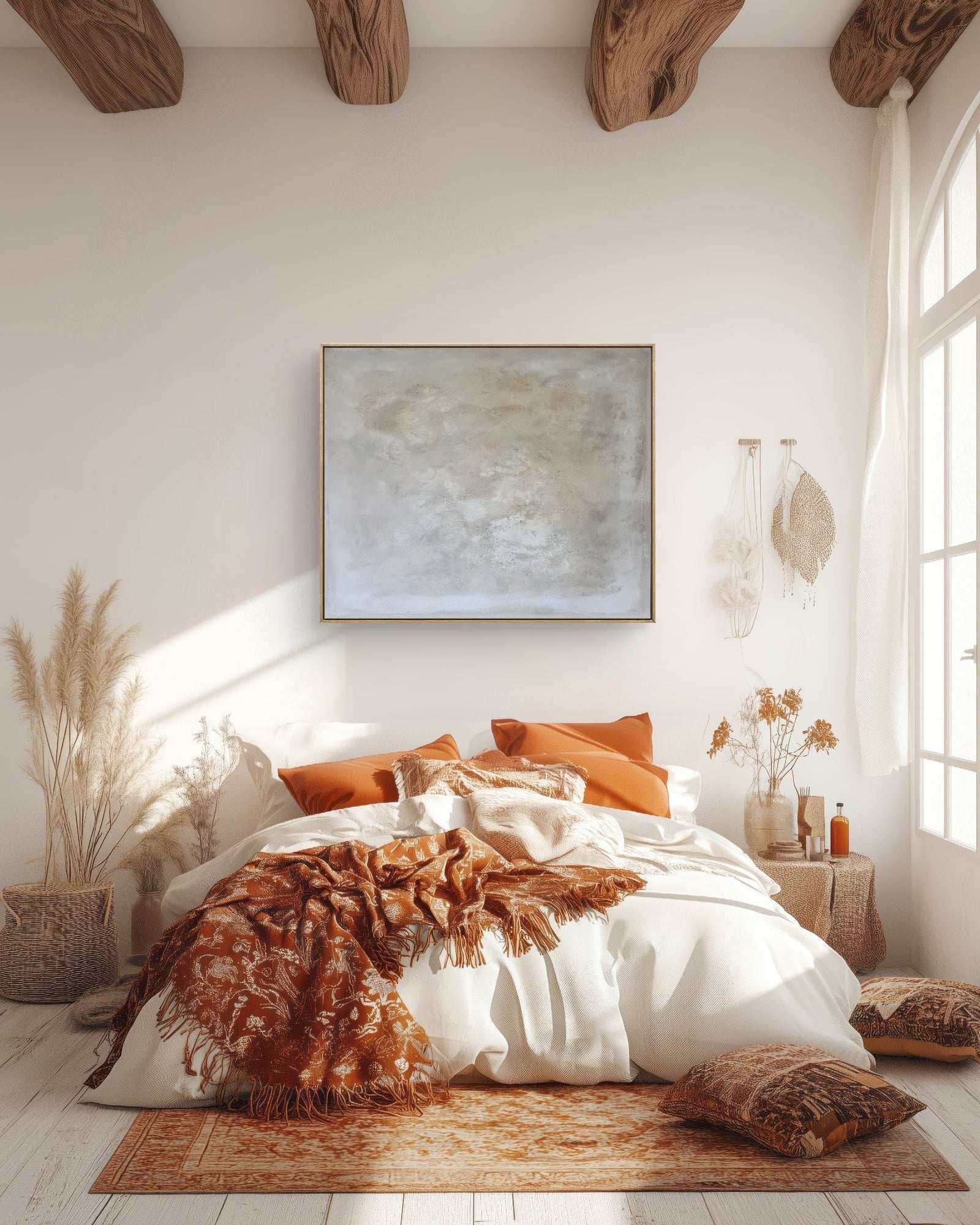 Sonnendurchflutetes, gemütliches Schlafzimmer mit weißer Bettwäsche, orange-braunen Kissen und Überwürfen, „Spurensuche“ von Conny Wachsmann über dem Bett in einem Schattenfugenrahmen, rustikalen Holzbalken, getrockneten Pflanzen, einem geflochtenen Korb und einem gemusterten Teppich auf hellem Holzboden.