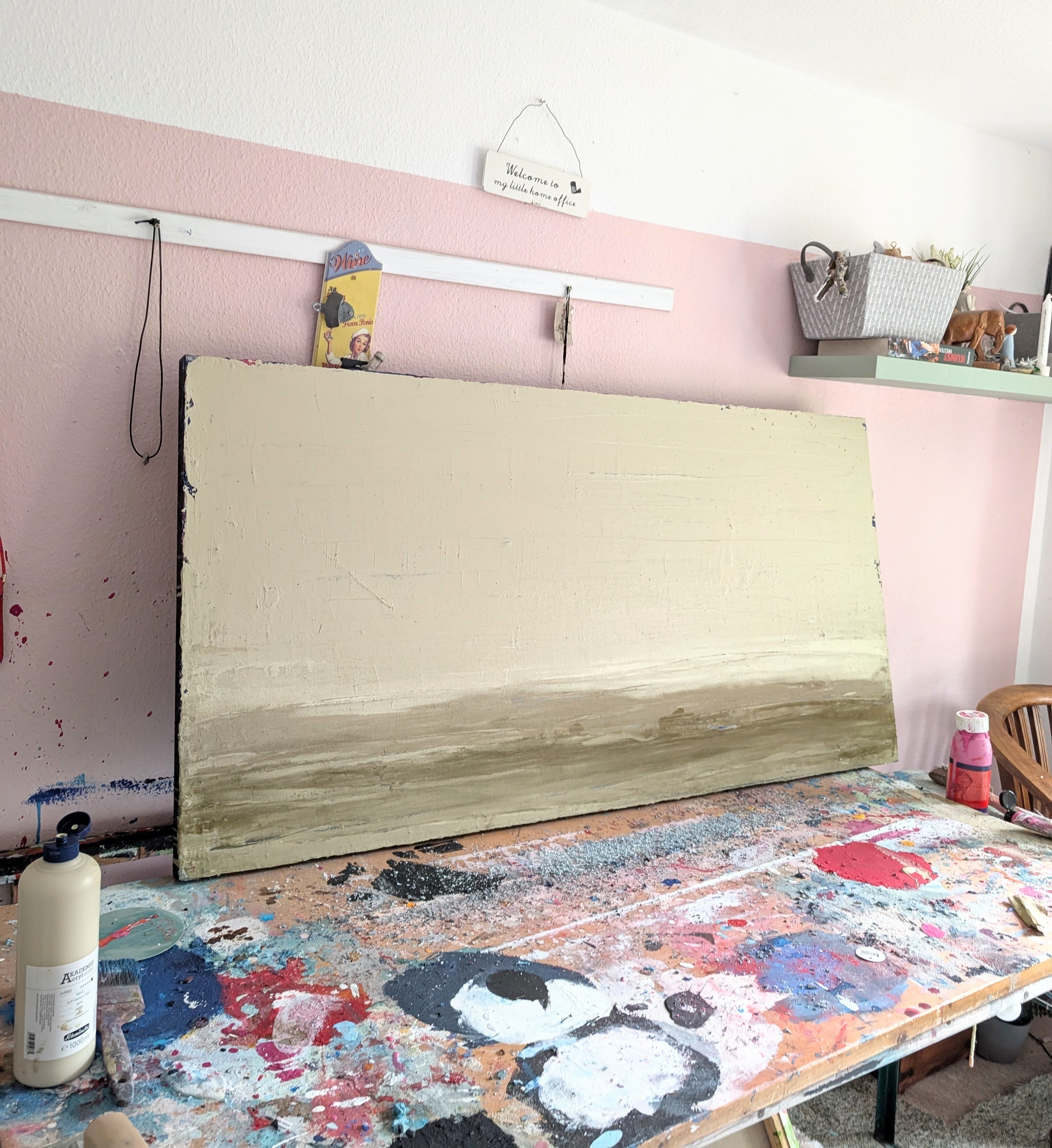 Zwischen Sommer und Herbst II", ein großes, unvollendetes Spachtelarbeit-Unikat von Conny Wachsmann, ruht auf einem bunten Arbeitstisch in einem hellen Atelier, das mit Regalen, Kunstzubehör und einem Korbsessel gefüllt ist.