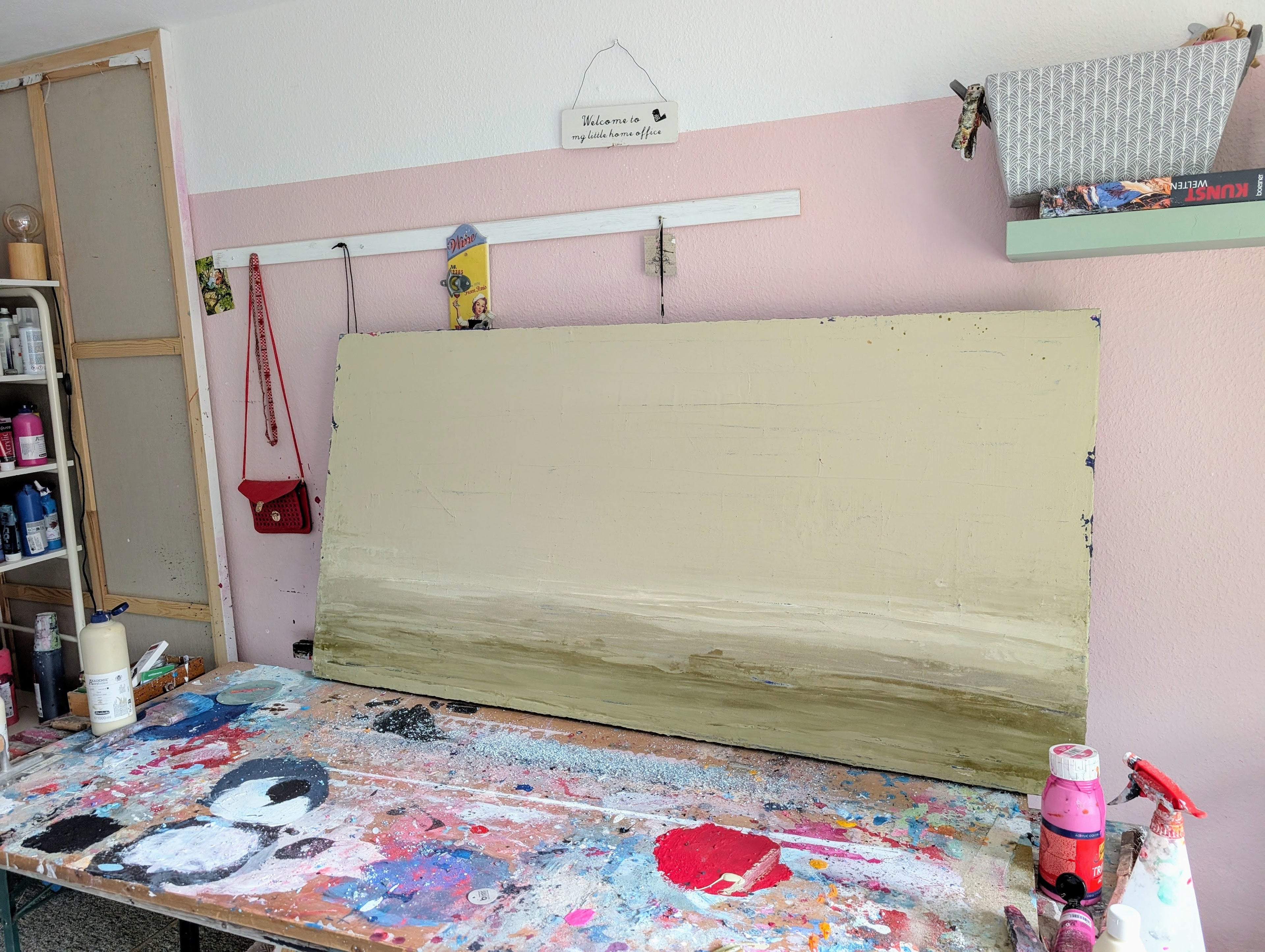 In einem unordentlichen Kunstatelier steht "Zwischen Sommer und Herbst II", ein Spachtelarbeit-Unikat von Conny Wachsmann. Das Kunstwerk lehnt an einer rosa-weißen Wand inmitten von verstreutem Kunstzubehör, Flaschen und Pinseln auf einem mit Farbe bespritzten Tisch. Darüber hängen ein Regal und ein Schild.