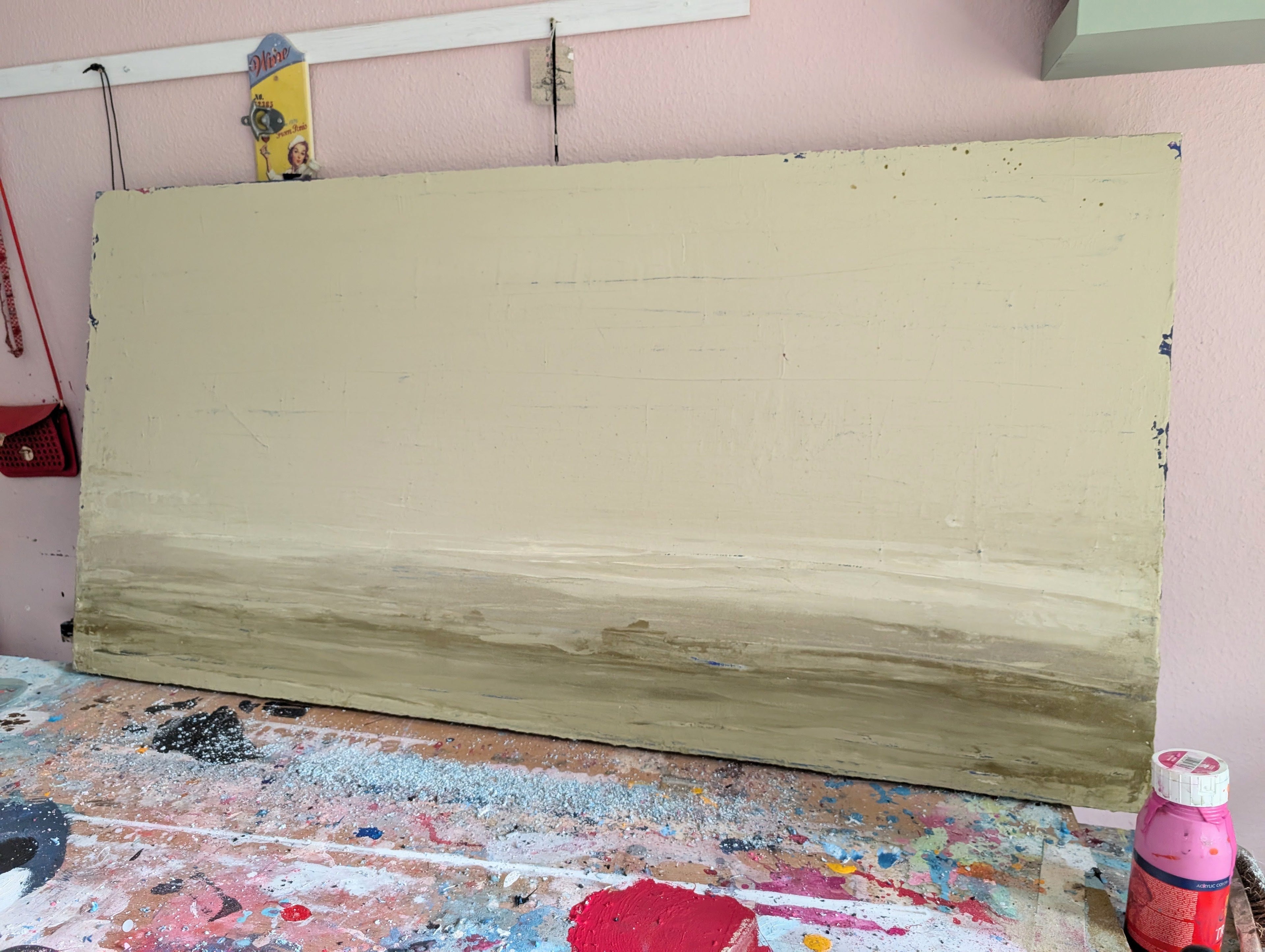 Zwischen Sommer und Herbst II von Conny Wachsmann, Künstlerin - eine große rechteckige Unikat-Spachtelarbeit in gedeckten Beige- und Brauntönen - steht auf einem mit Farbe bespritzten Tisch neben Kunstzubehör vor einer rosafarbenen Wand, die dieses auffällige Originalgemälde präsentiert.