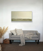 Ein hellgraues Sofa mit strukturierten Kissen und einer Strickdecke steht vor einer weißen Wand, darunter "Zwischen Sommer und Herbst II" von Conny Wachsmann. Ein Korb und Pampasgras-Dekor stehen links auf einem braunen Teppichboden.