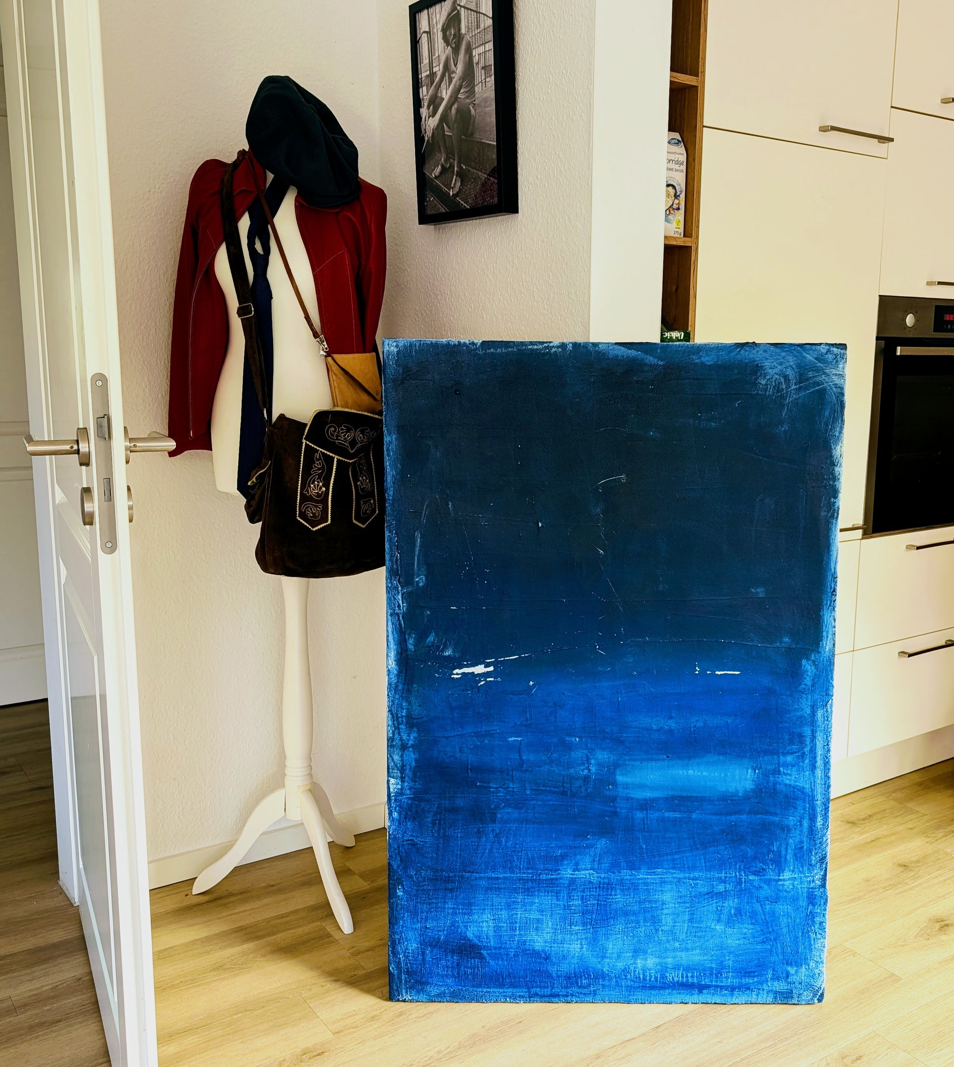 Ein großes "Fjord" Acryl auf Leinwand von Conny Wachsmann, ein signiertes Unikat in Blau, lehnt an der Küchenwand; daneben steht eine weiße Schaufensterpuppe in Rot an der offenen Tür unter einem Schwarz-Weiß-Foto.